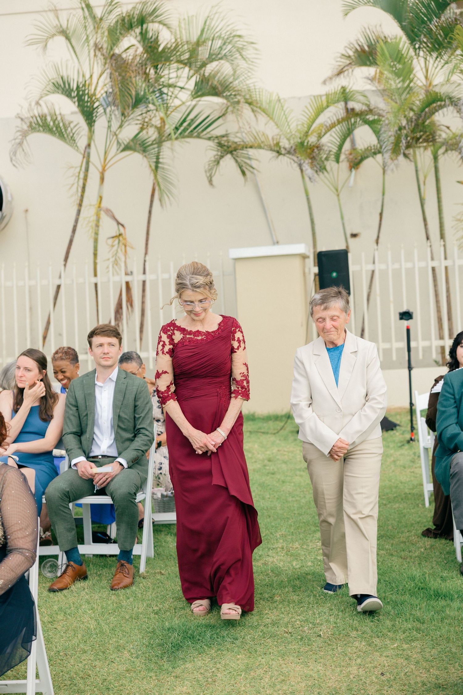 Wedding guests seated on white chairs on a lawn with palm trees in the background during an outdoor ceremony.