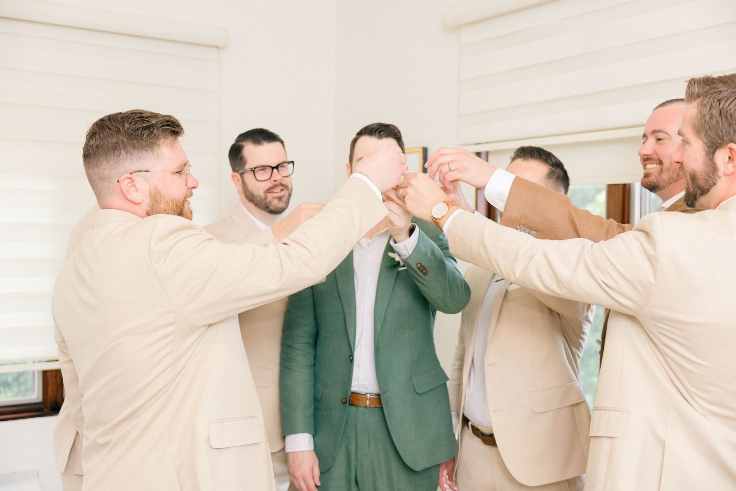Wedding party in beige and green suits celebrating with raised arms in bright indoor space.
