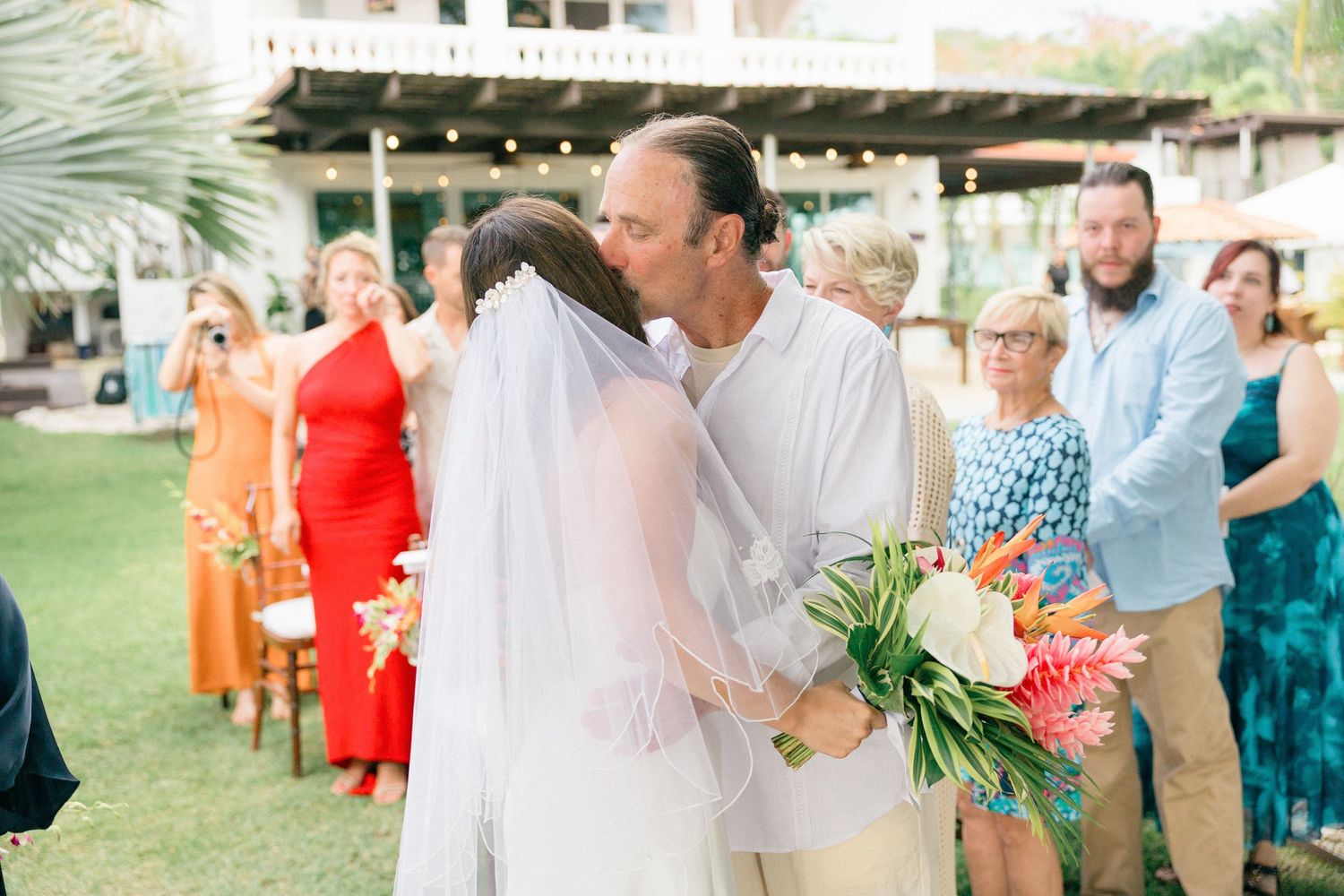 Wedding guests mingle on lawn with tropical decor and bright flowers.