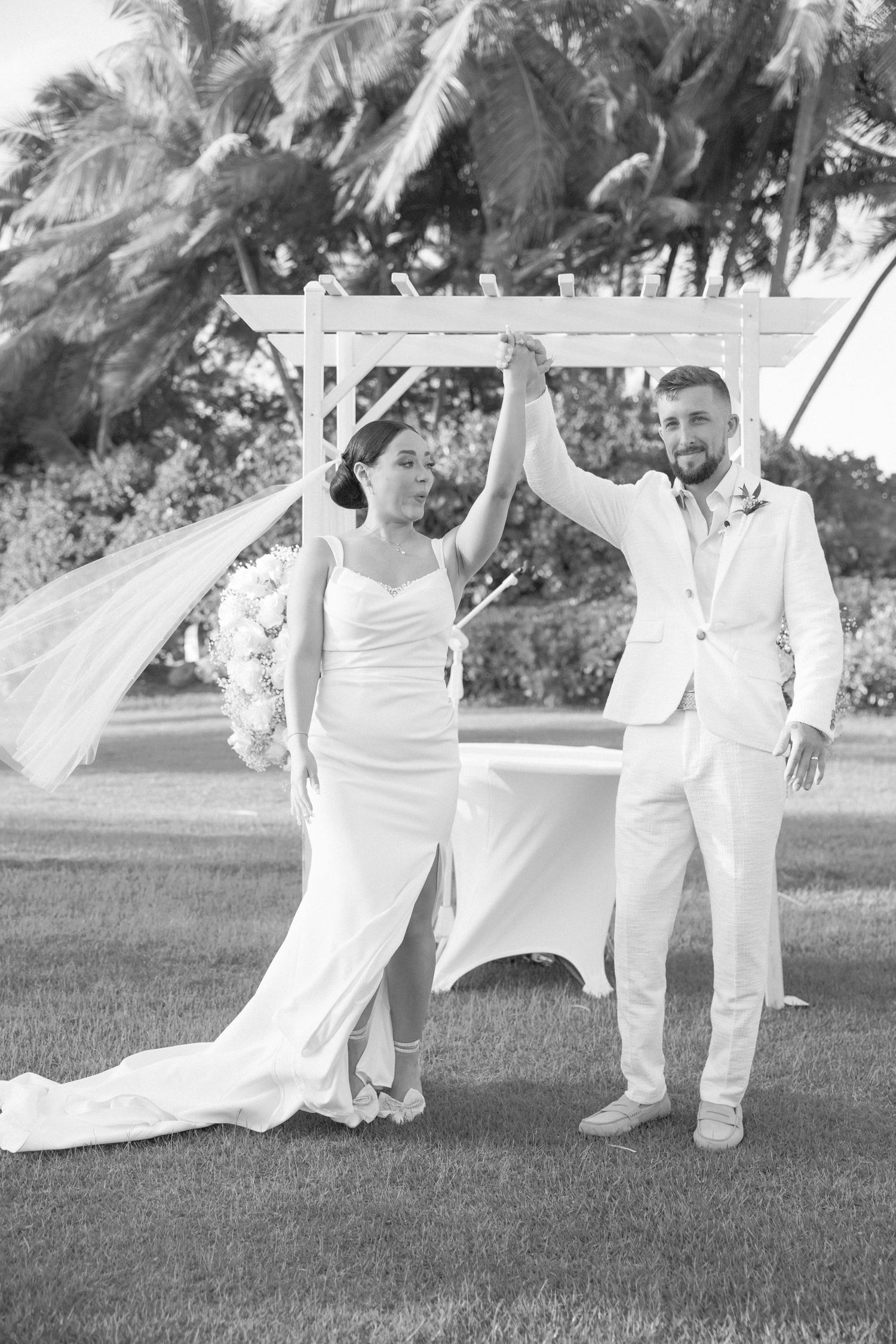 Wedding party dances with white fabric panels on a grassy lawn surrounded by palm trees.