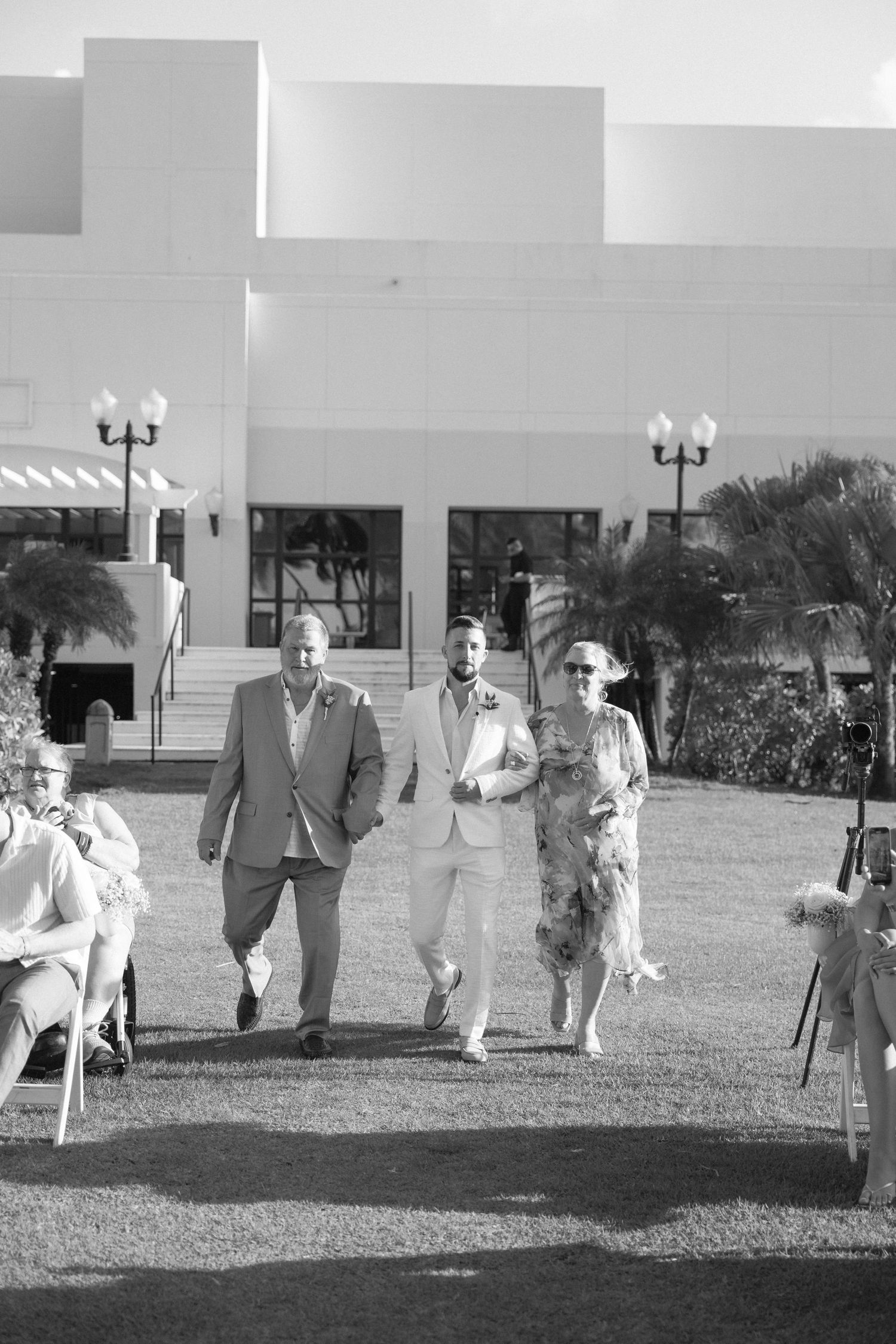 Black and white photo of wedding party walking across a grassy lawn near a modern building.