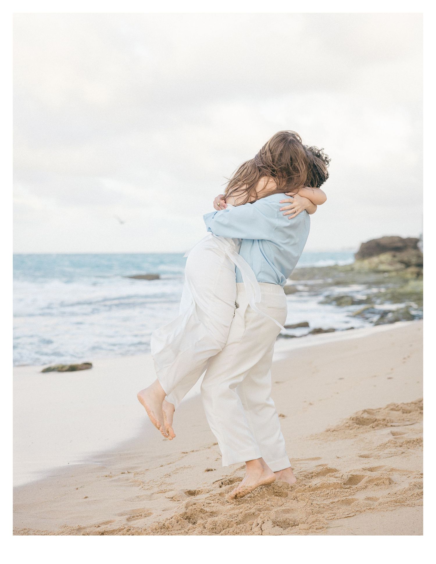 Couple walking barefoot together on a sandy beach at sunset wearing white and light blue casual outfits.