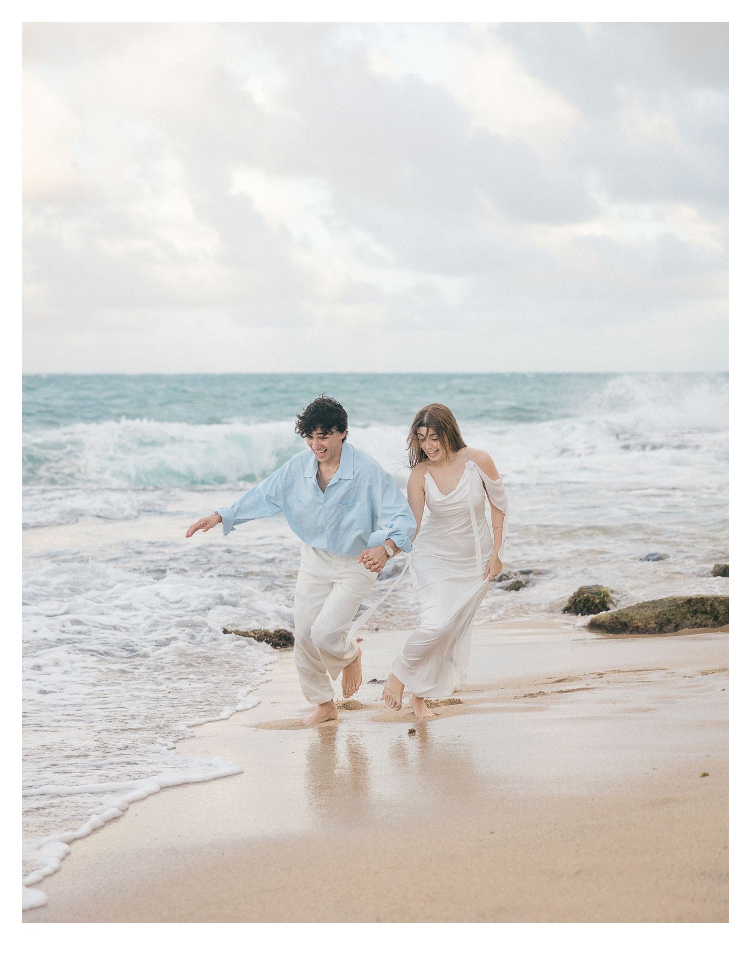 Couple in white and blue outfits walking together along rocky beach shoreline.