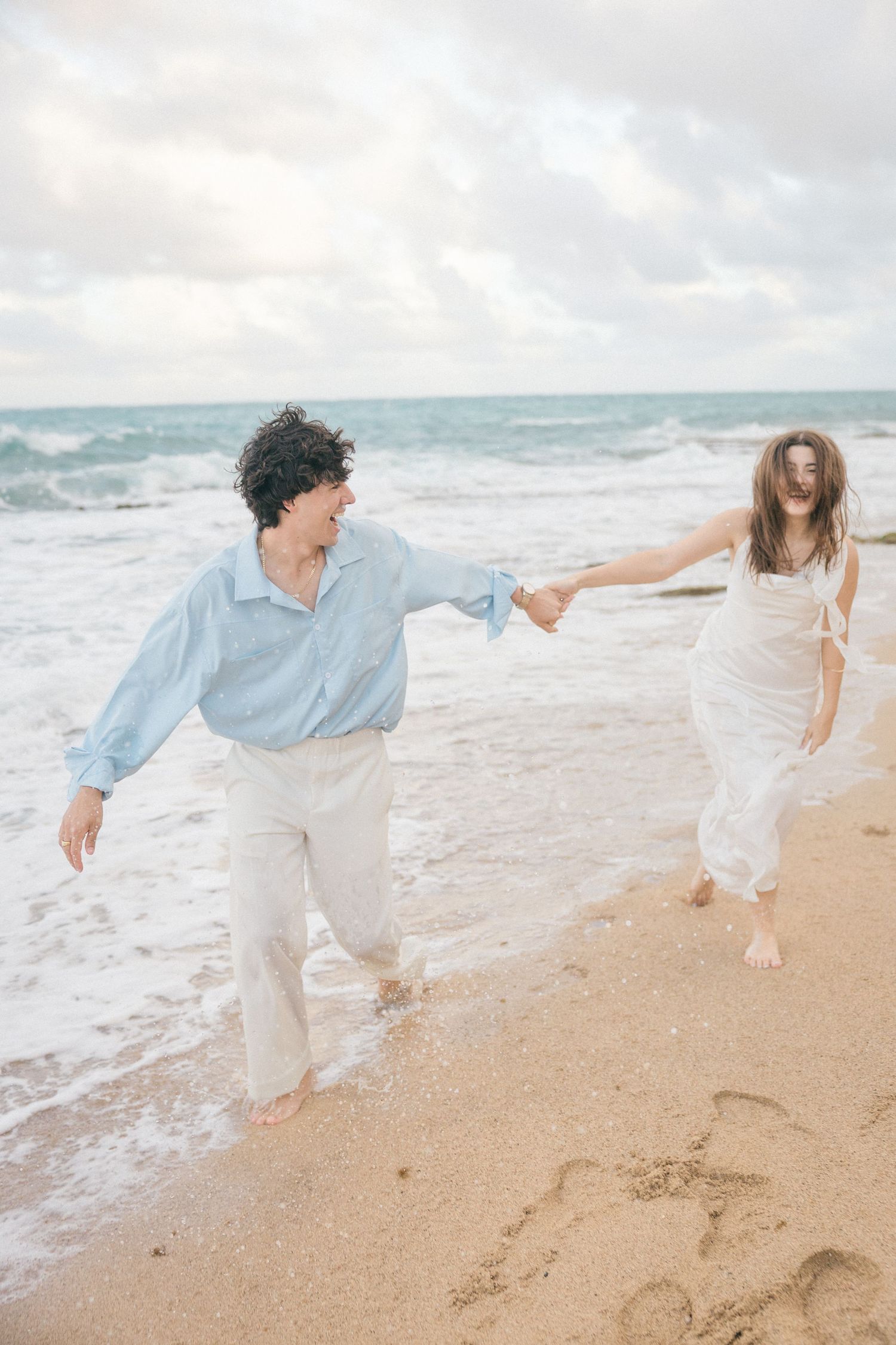 Two people dance and play together on a beach with waves crashing behind them.