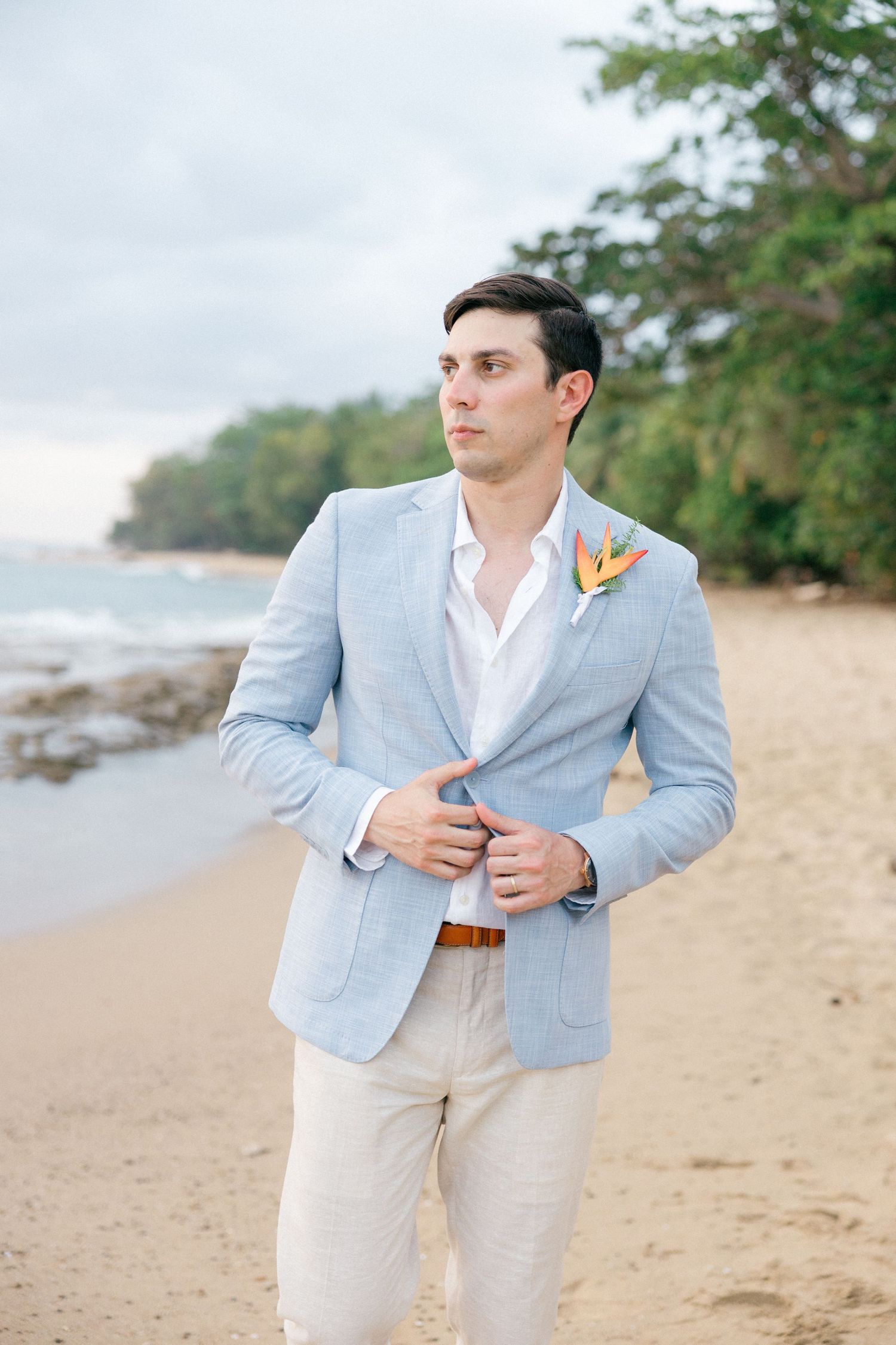 Groom in light blue blazer and cream pants poses on beach with boutonniere.