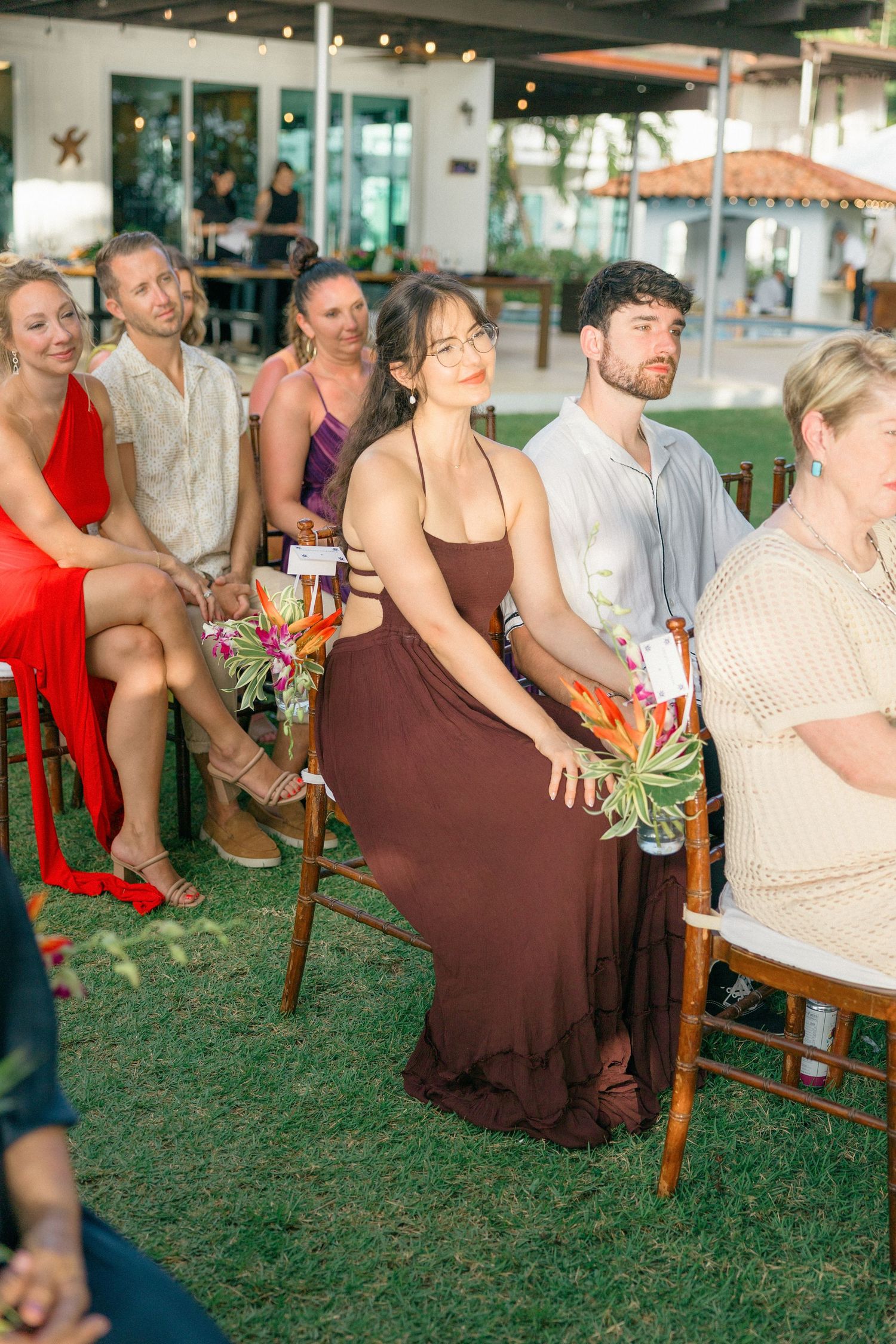 Wedding guests seated in wooden chairs on grass watching a ceremony.