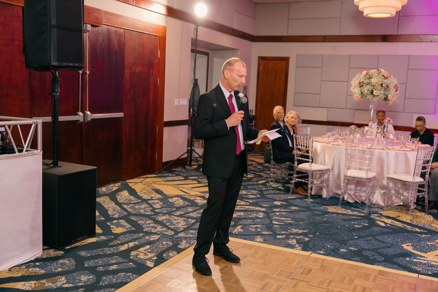 Speaker addresses wedding guests from dance floor in reception ballroom with blue carpet.