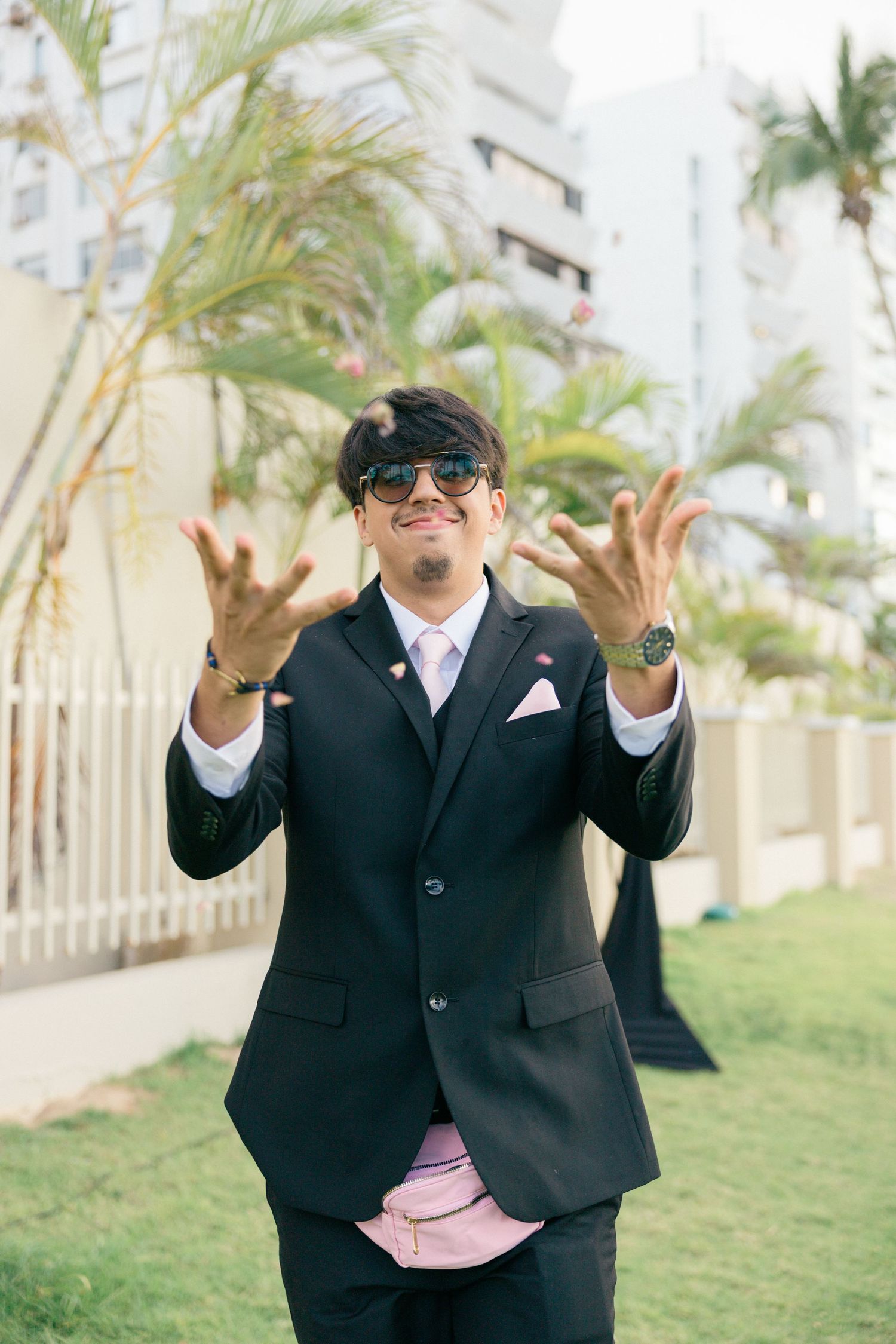 Wedding guest in sunglasses and suit gestures playfully on grass.