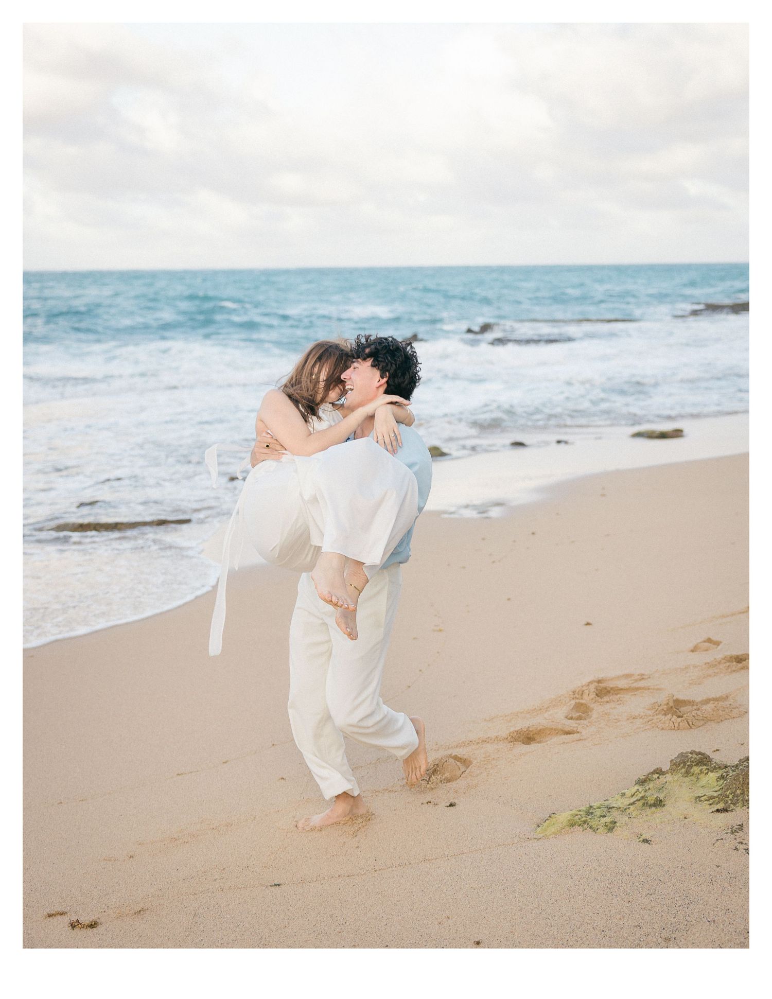 A couple embracing on a sandy beach with ocean waves in the background.