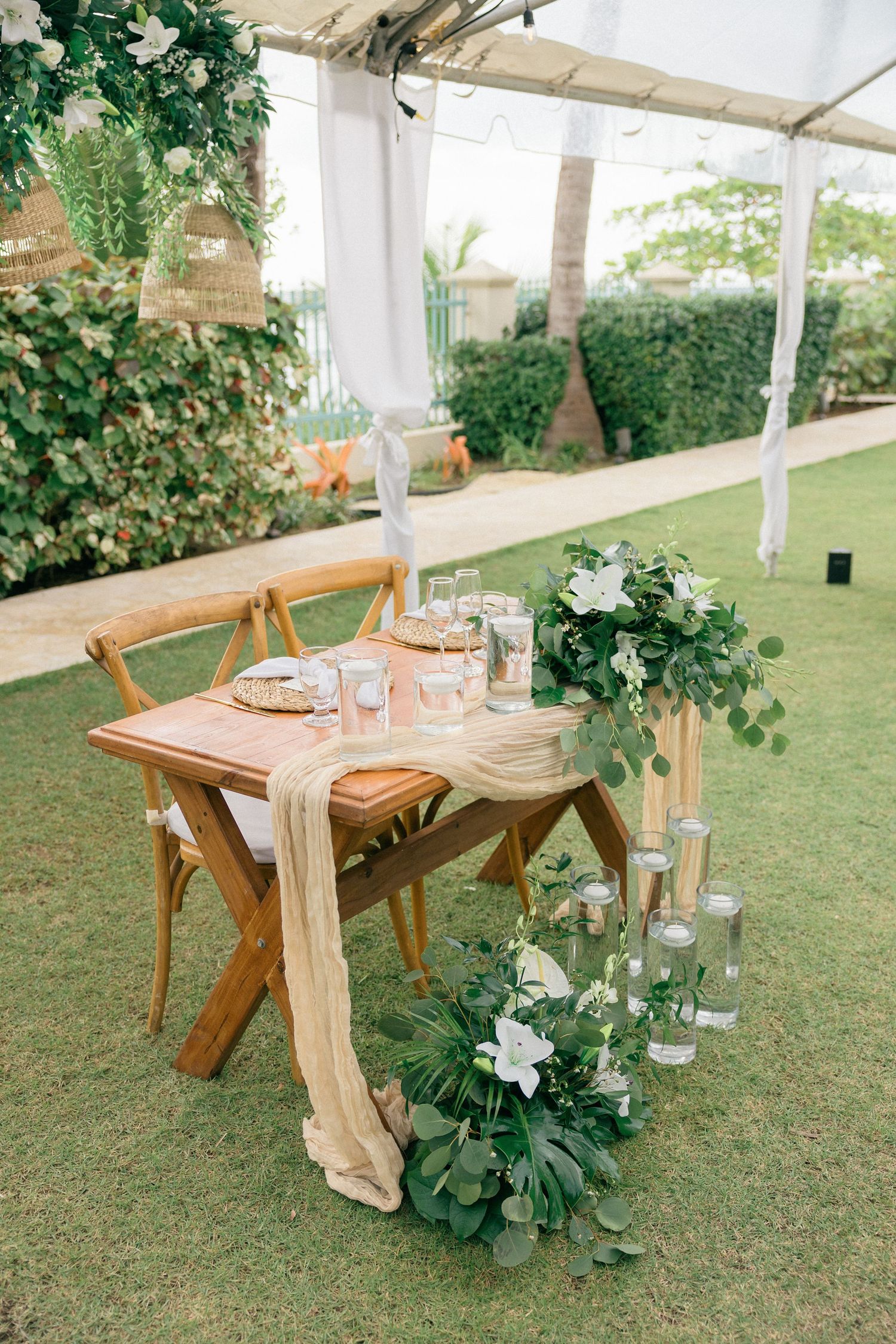 Wooden farm tables set for outdoor reception under white tent with natural decor.
