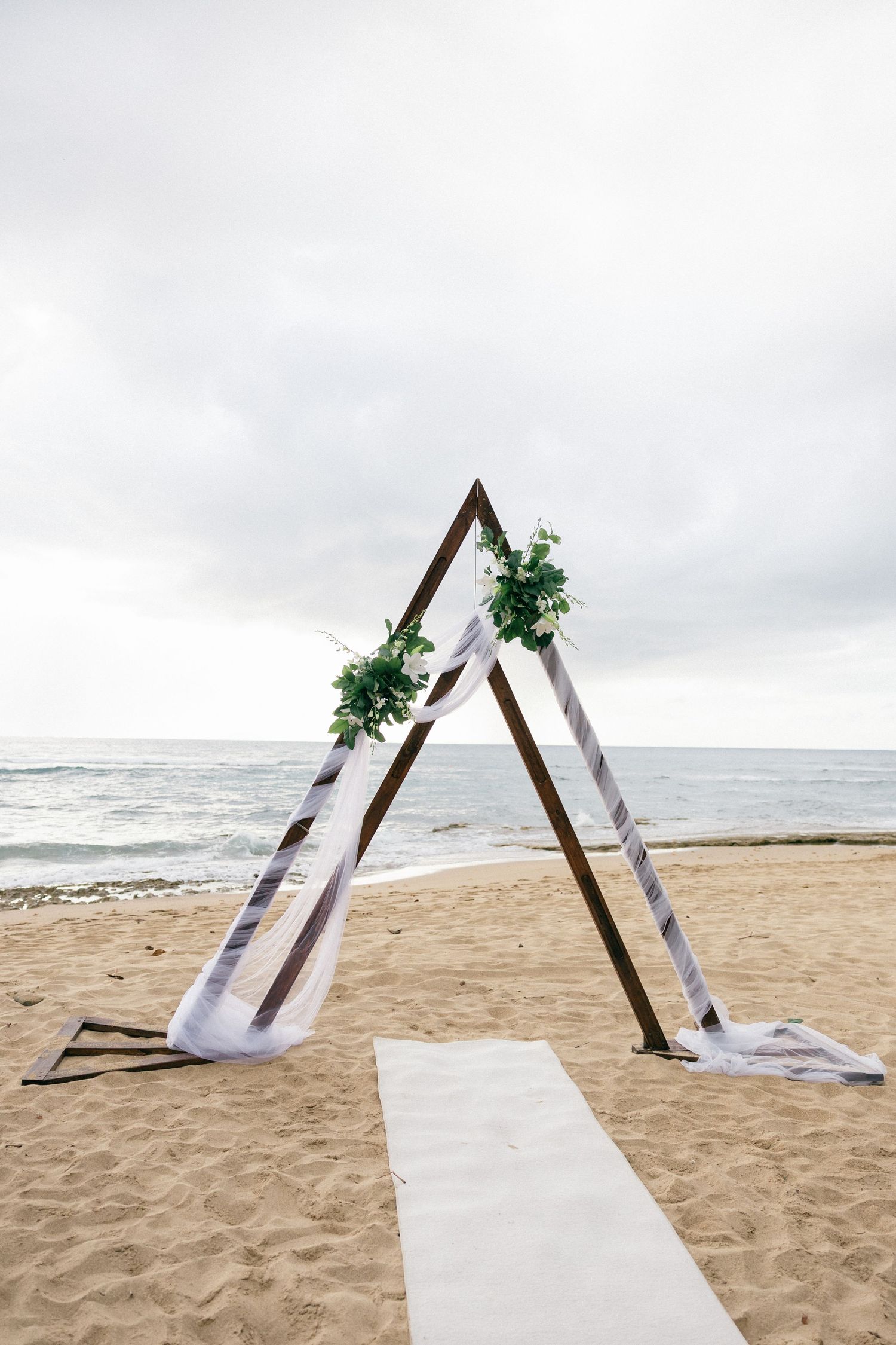 Minimalist wooden triangle arch setup on a beach with white fabric draping and greenery accents.