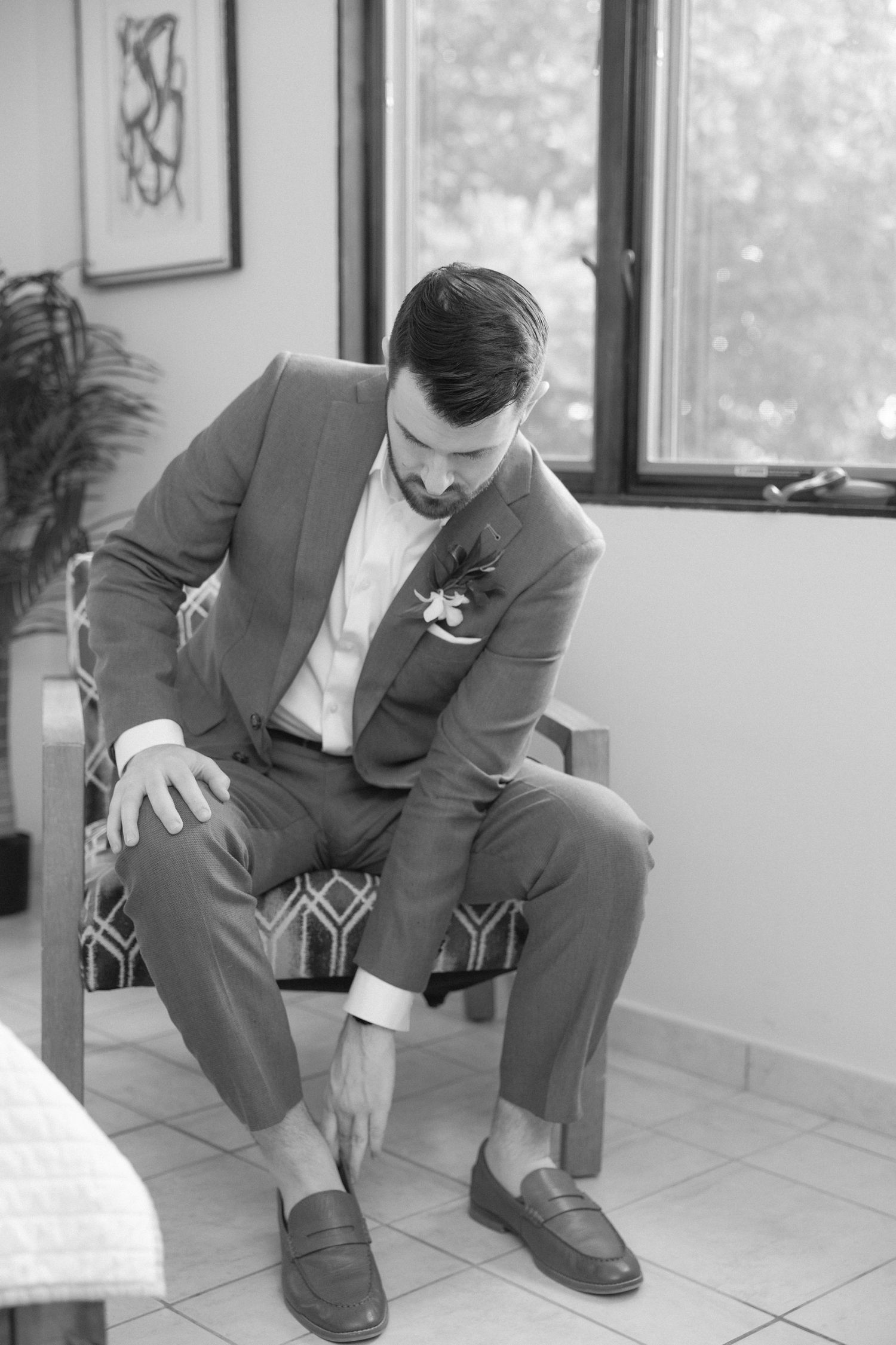 Black and white portrait of groom seated in modern chair wearing formal attire.