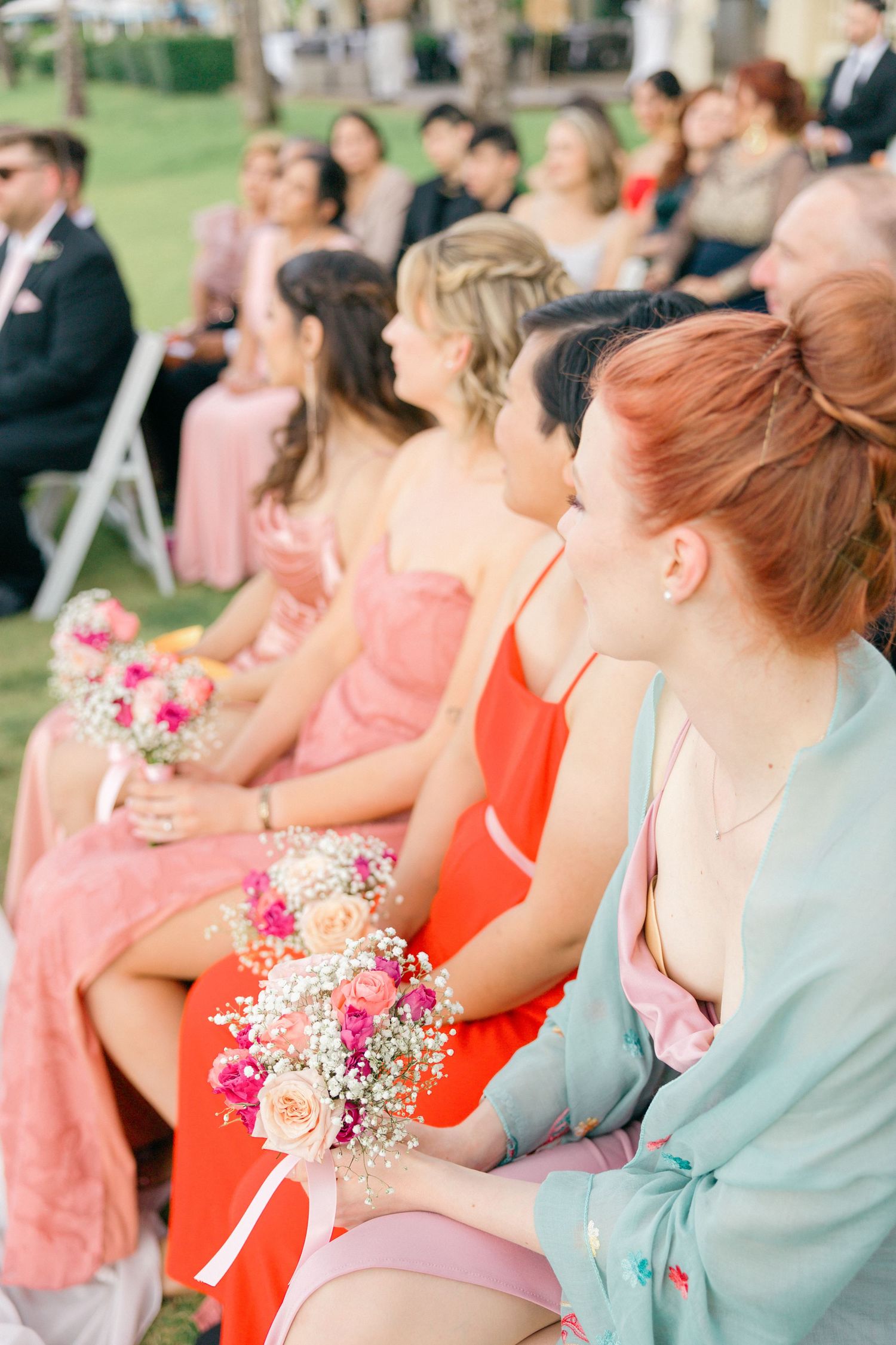 Wedding party members in pastel dresses hold small pink and white bouquets during ceremony.