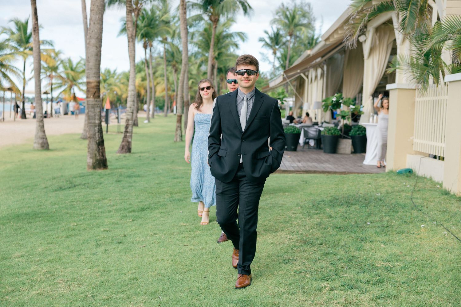 Wedding guest in dark suit walks along palm tree-lined path at tropical venue.