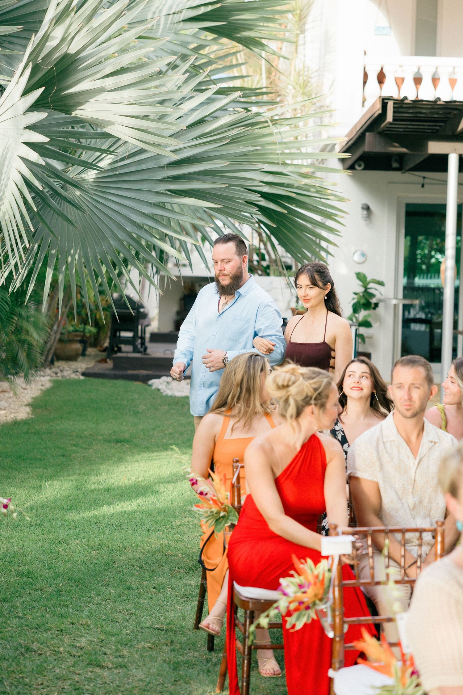 Outdoor wedding ceremony setup with palm fronds and guests in bright summer attire.