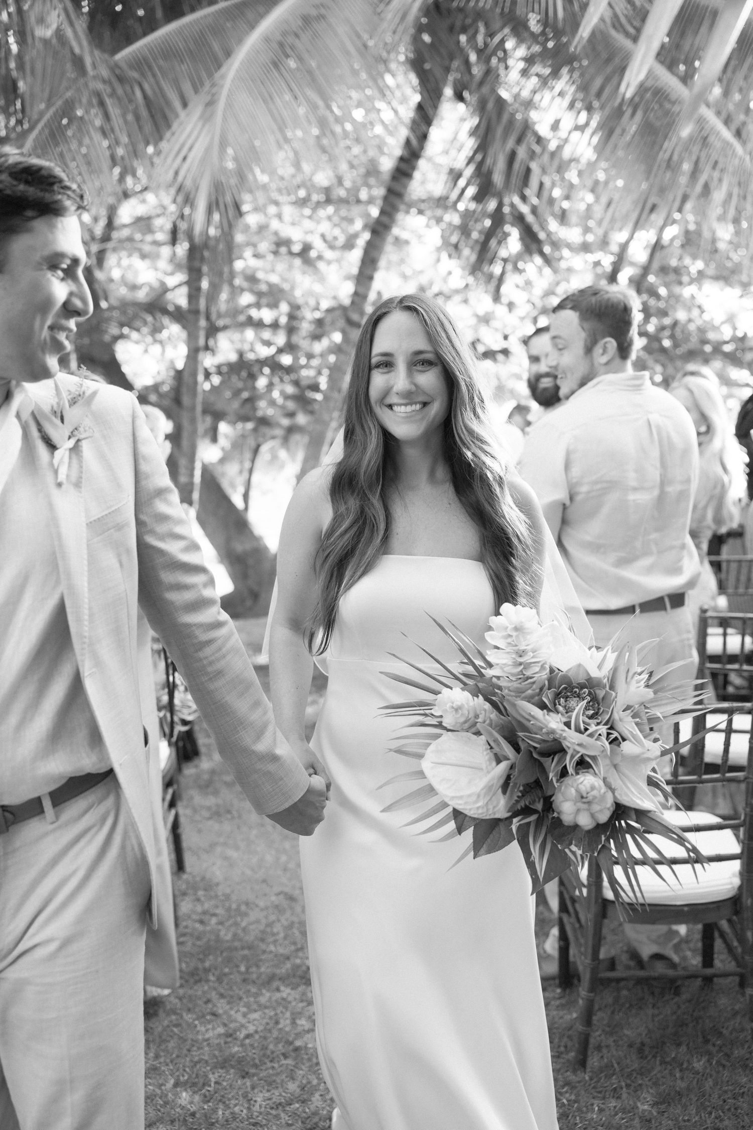 Wedding party in white and light colored attire posing outdoors with tropical bouquets.