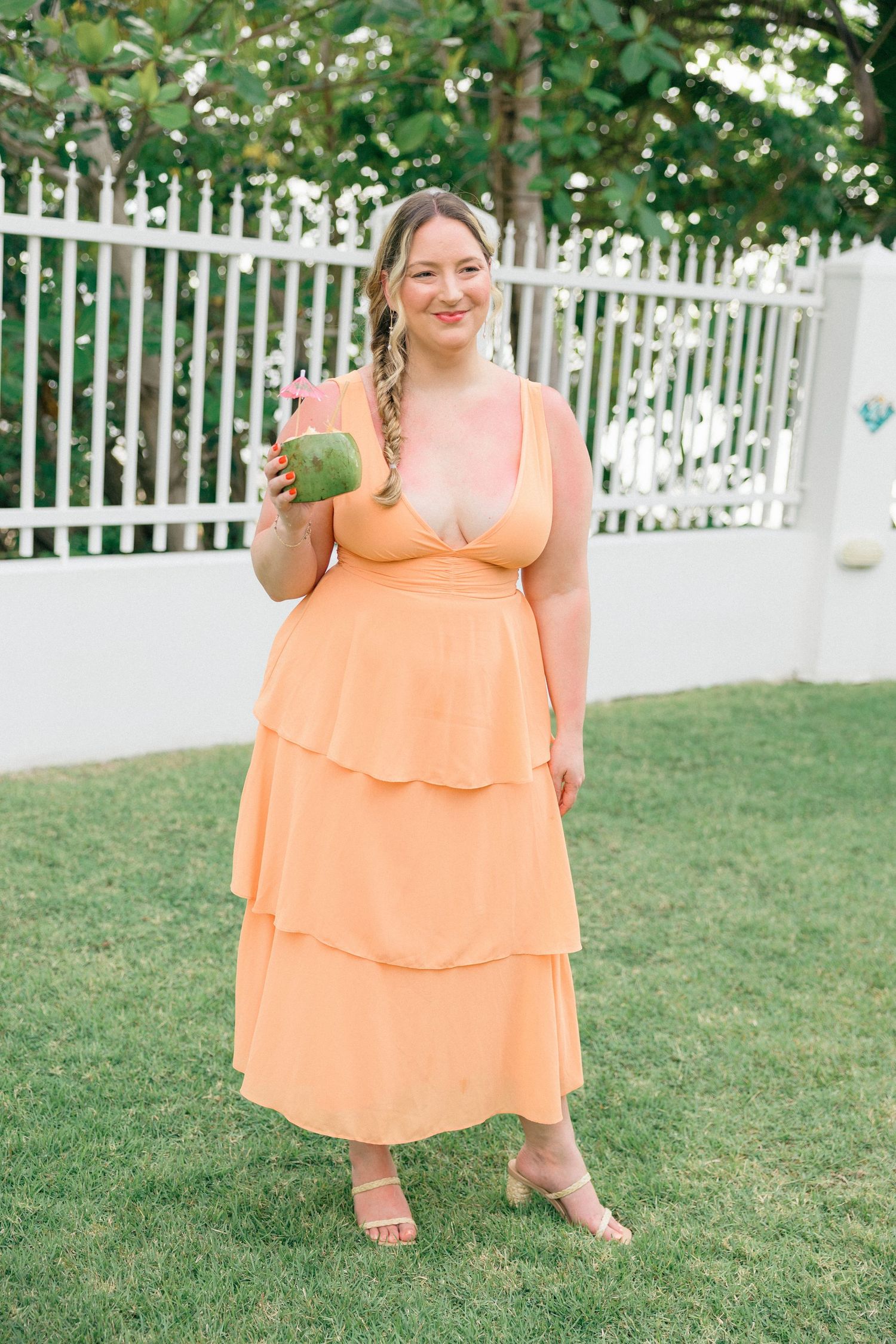 A person in a peach-colored tiered dress holding a coconut drink in a garden setting.