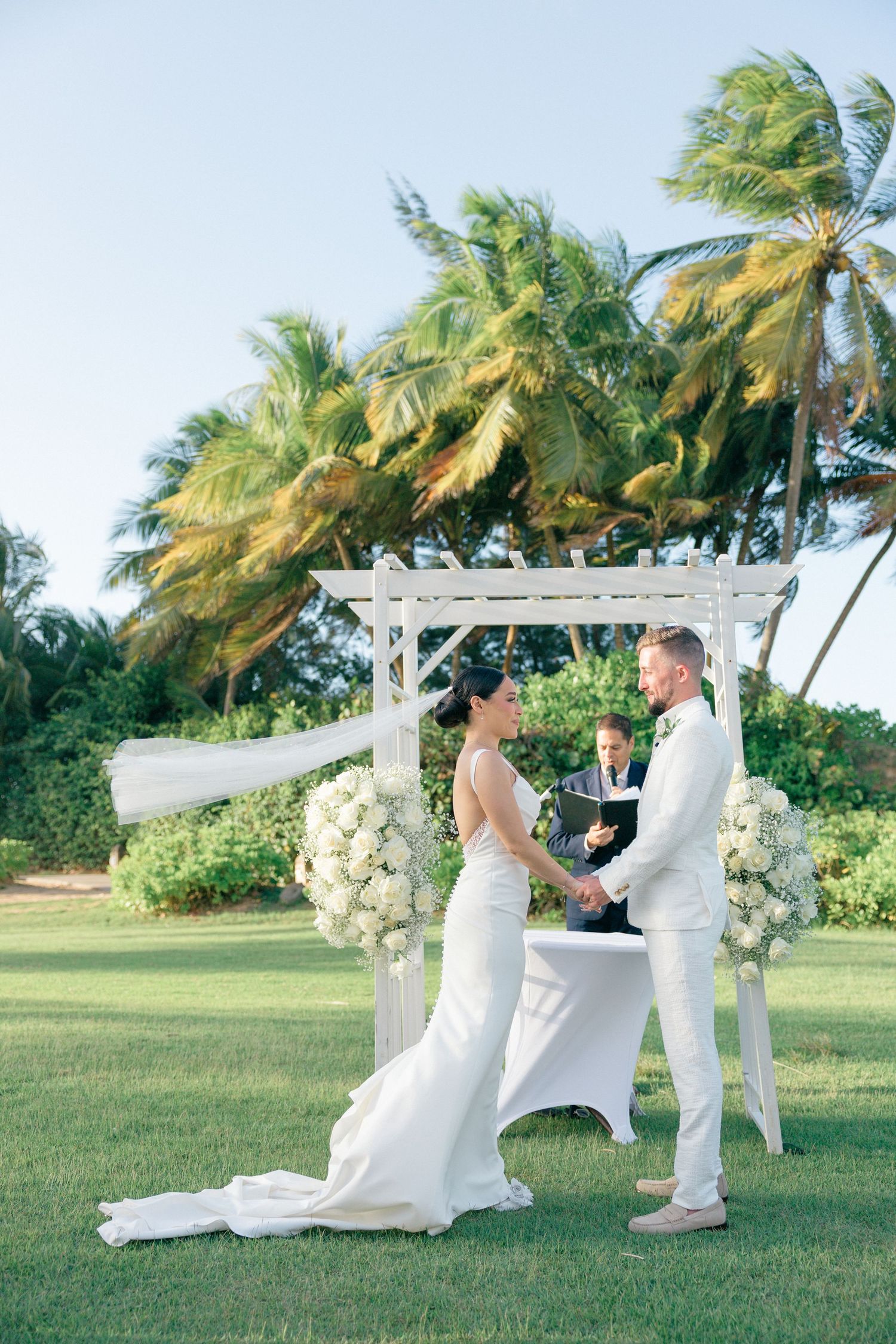 Intimate wedding ceremony moment under a white pergola with tropical landscaping.