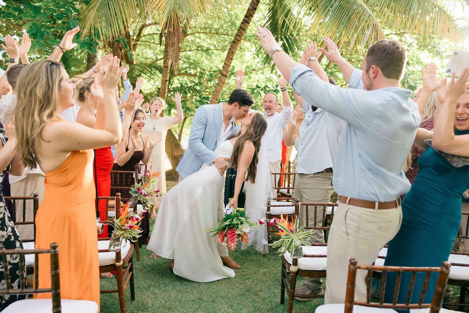 Outdoor wedding reception setup with wooden chairs under palm trees and string lights.