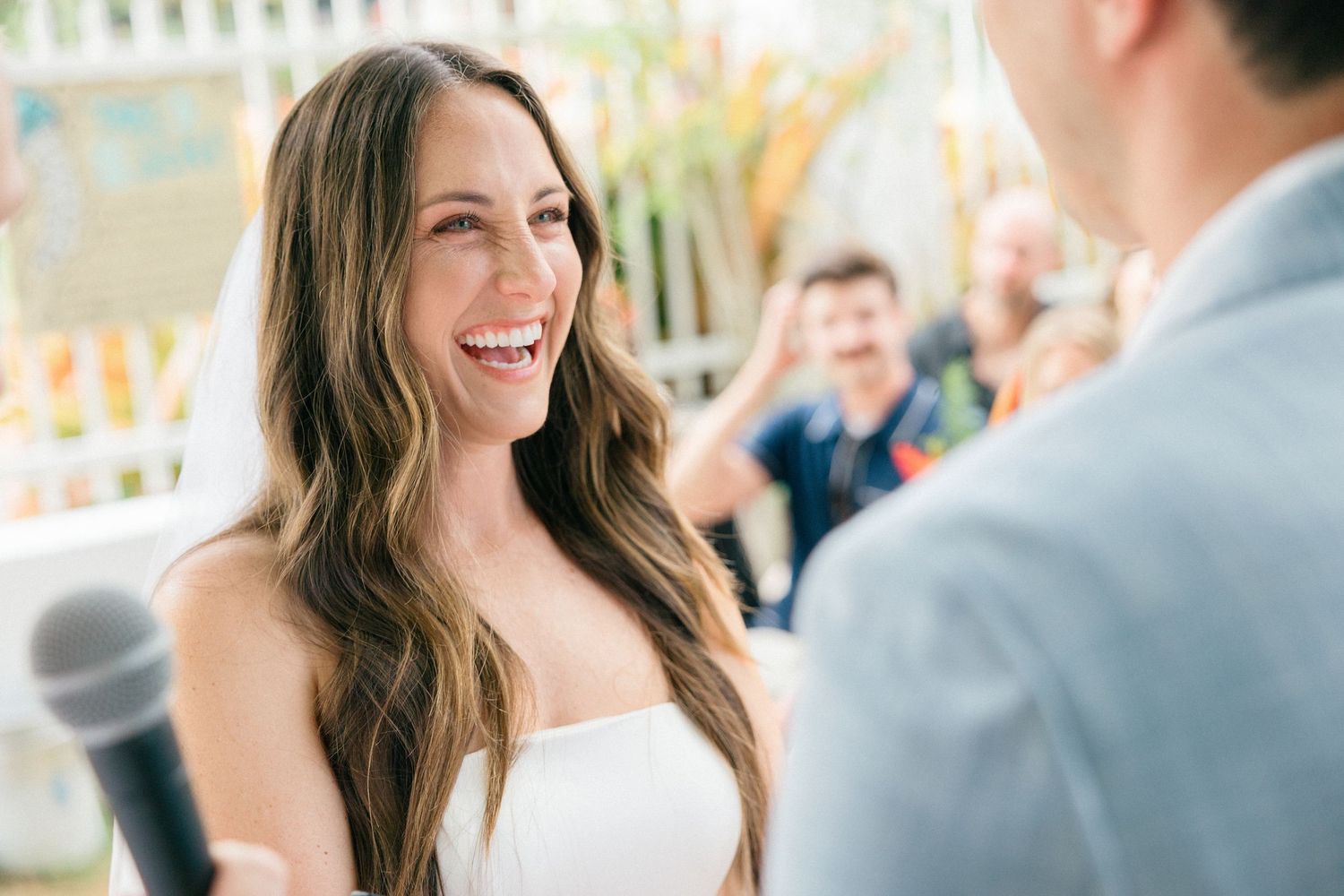 Bride in white strapless wedding dress smiles joyfully during outdoor ceremony.