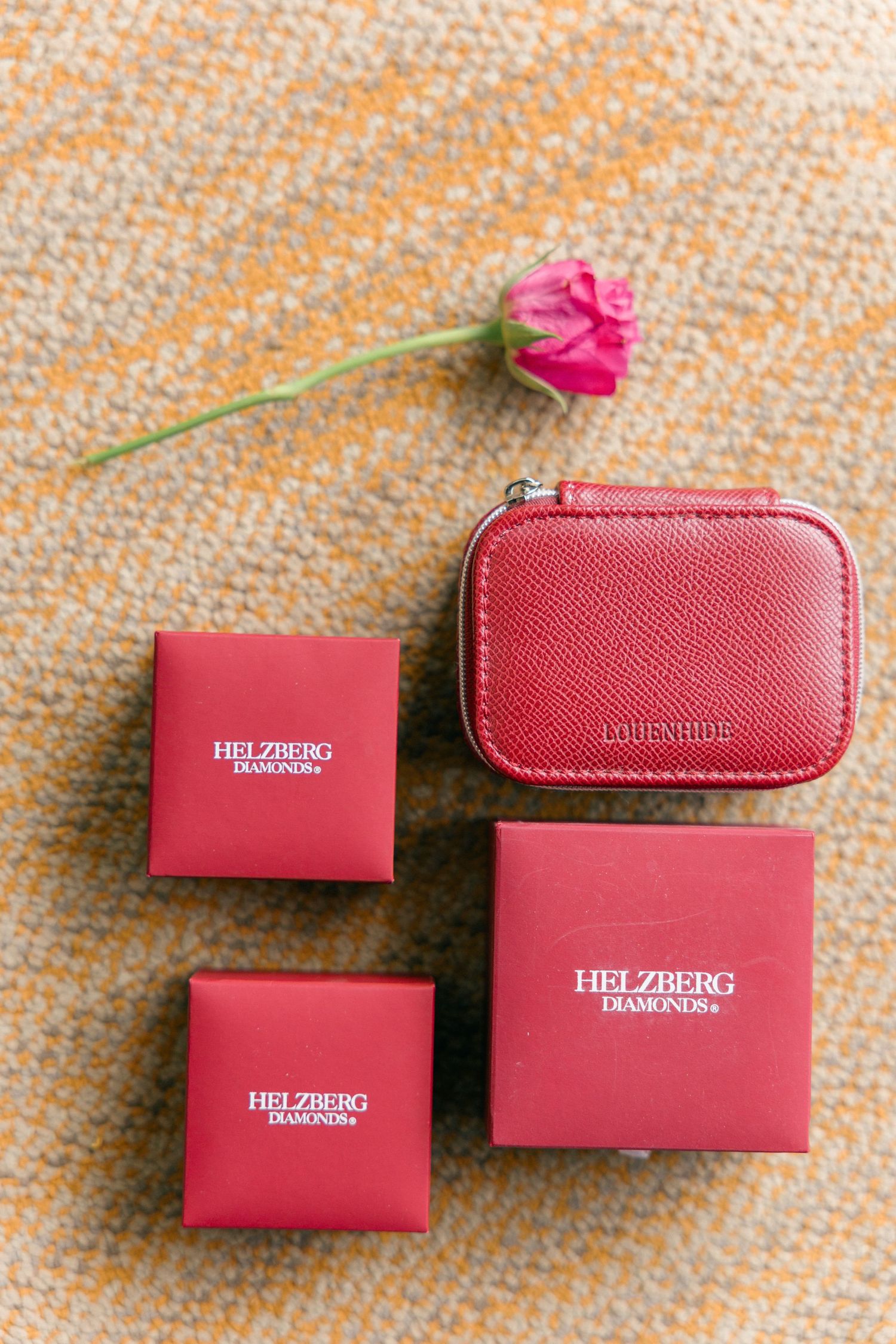 Pink leather accessories and gift boxes arranged with a single rose on textured fabric.