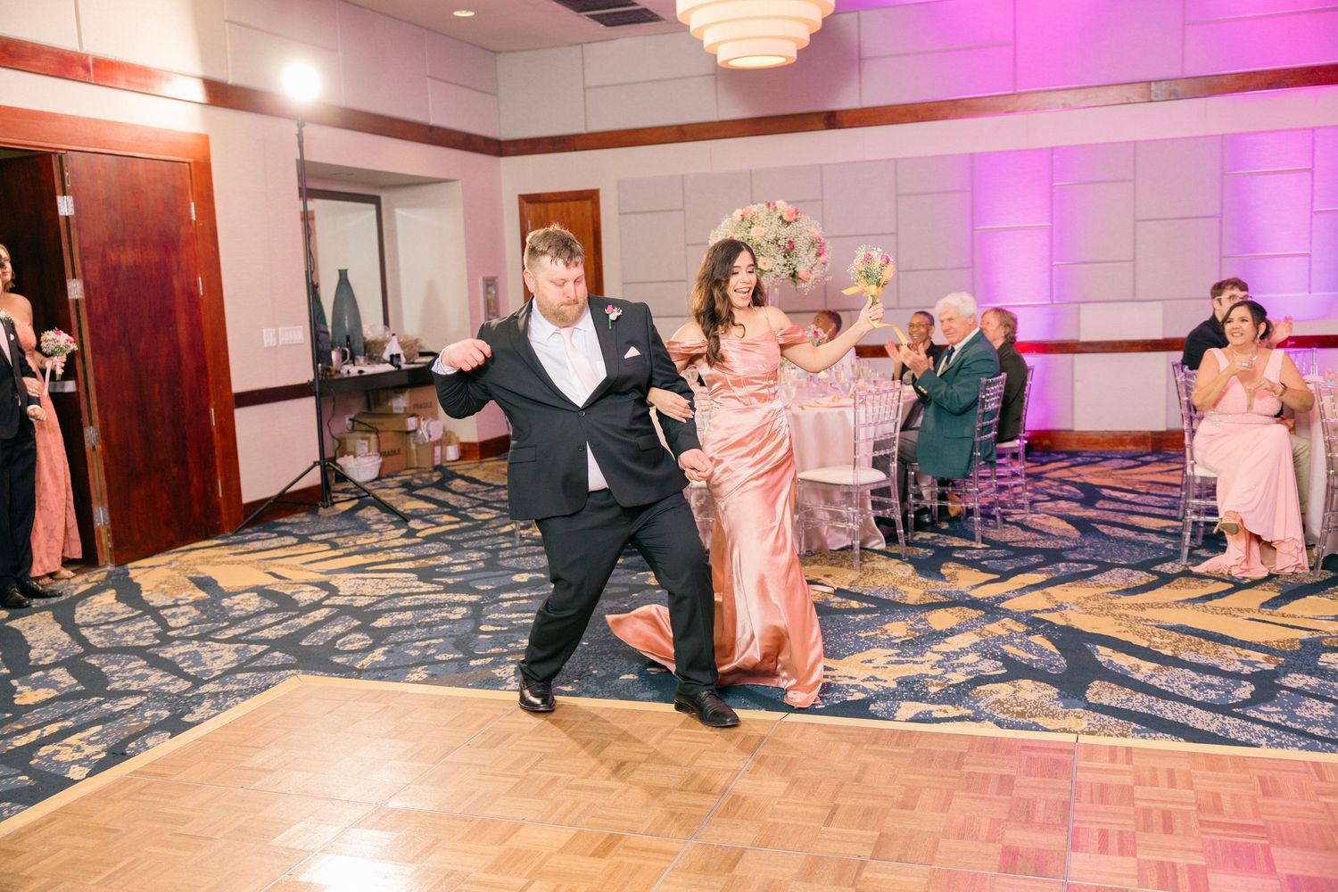 Couple dances energetically in reception hall with pink lighting and chandelier.