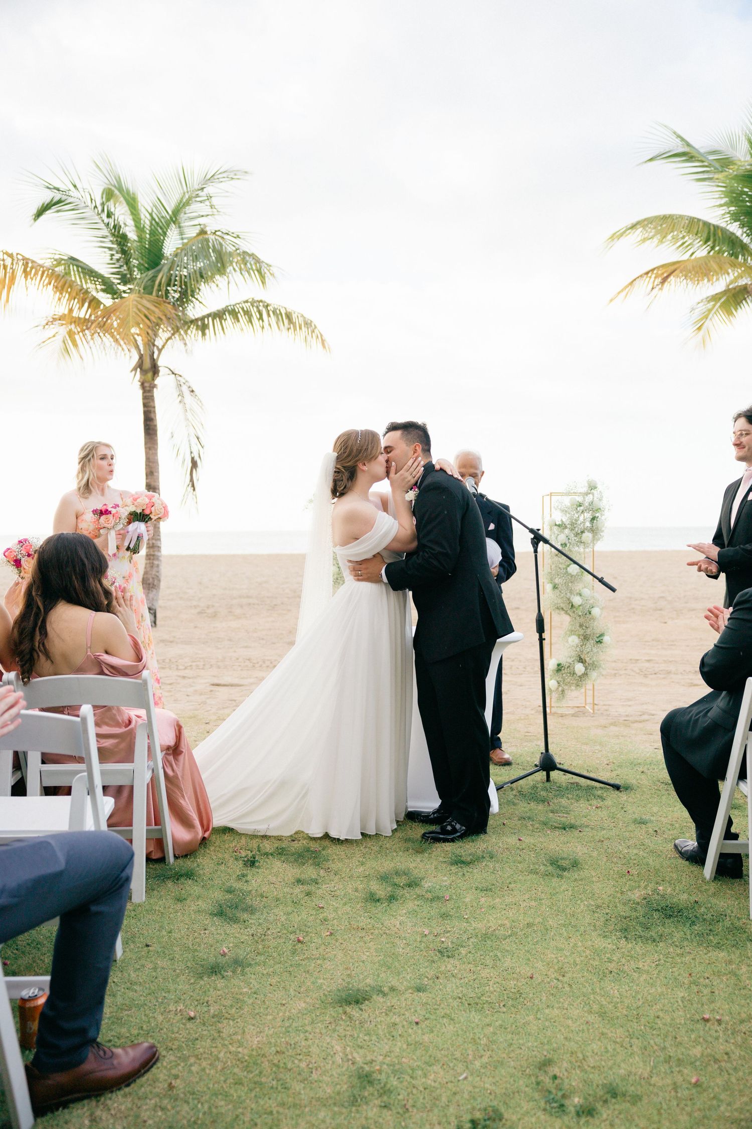Outdoor wedding ceremony setup with white chairs and palm trees along a beachfront.
