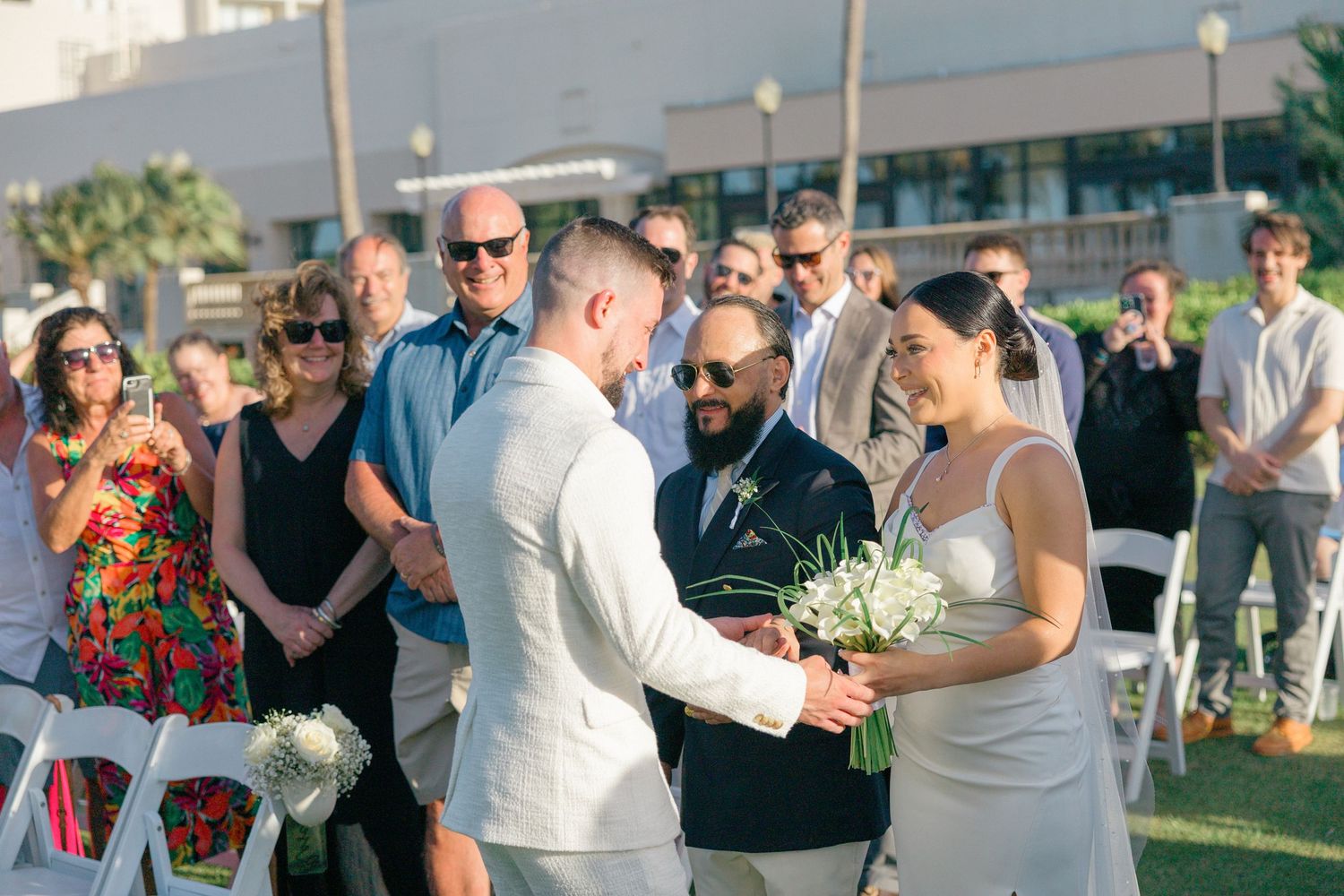 Wedding guests gather outdoors for a celebration with palm trees and modern architecture in the background.