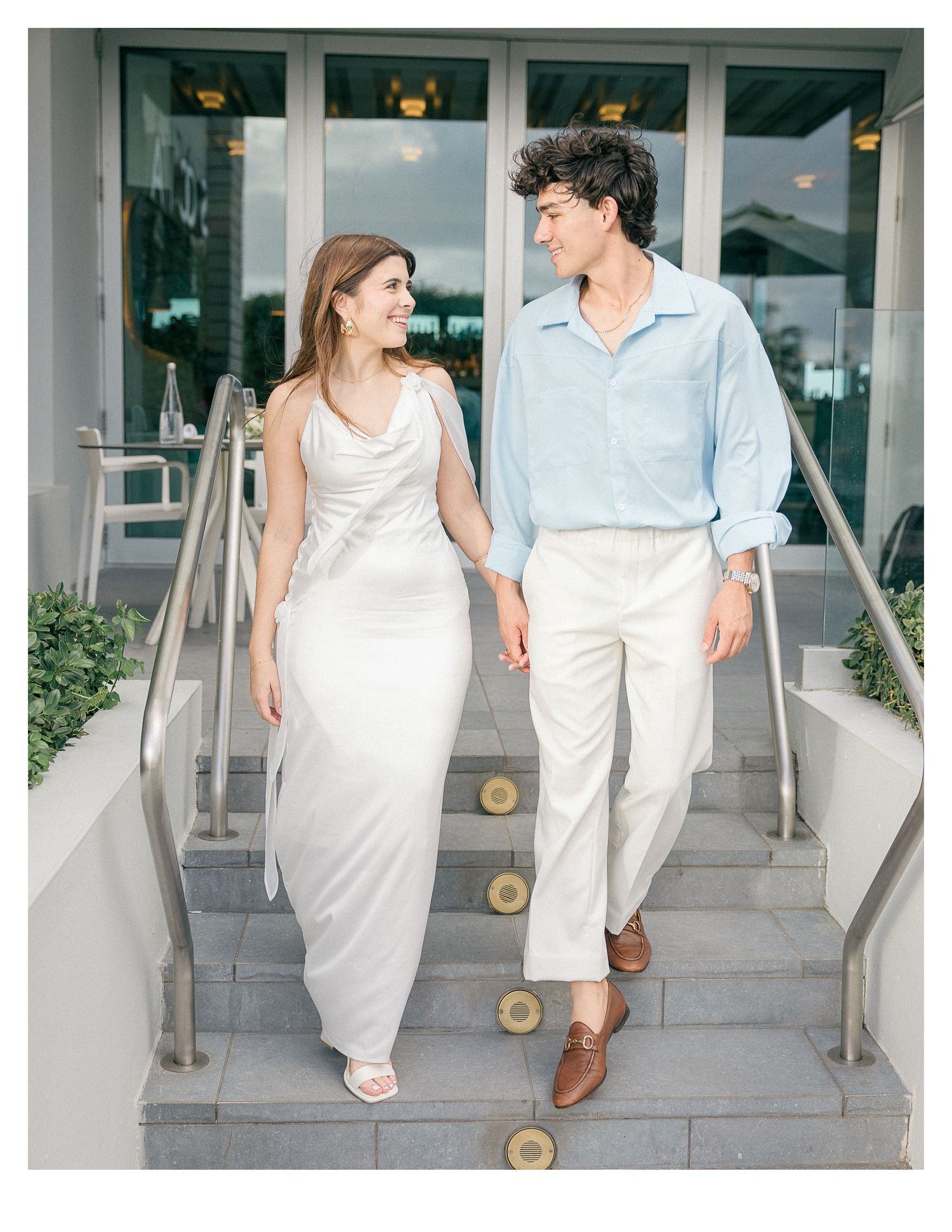 A couple in light summer attire walks down concrete steps together holding hands.