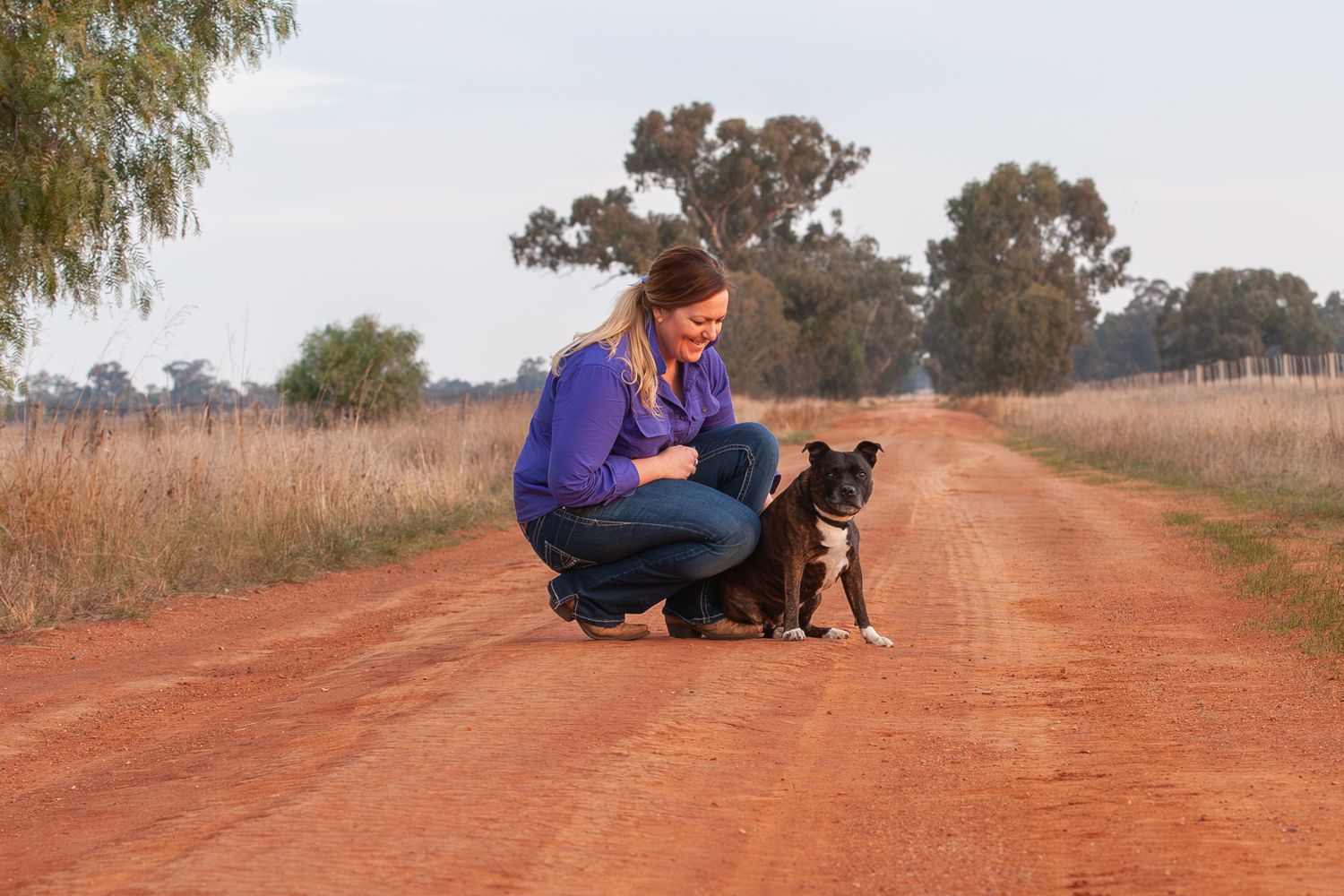 Person in purple shirt crouches next to a small black dog on a rural dirt road.