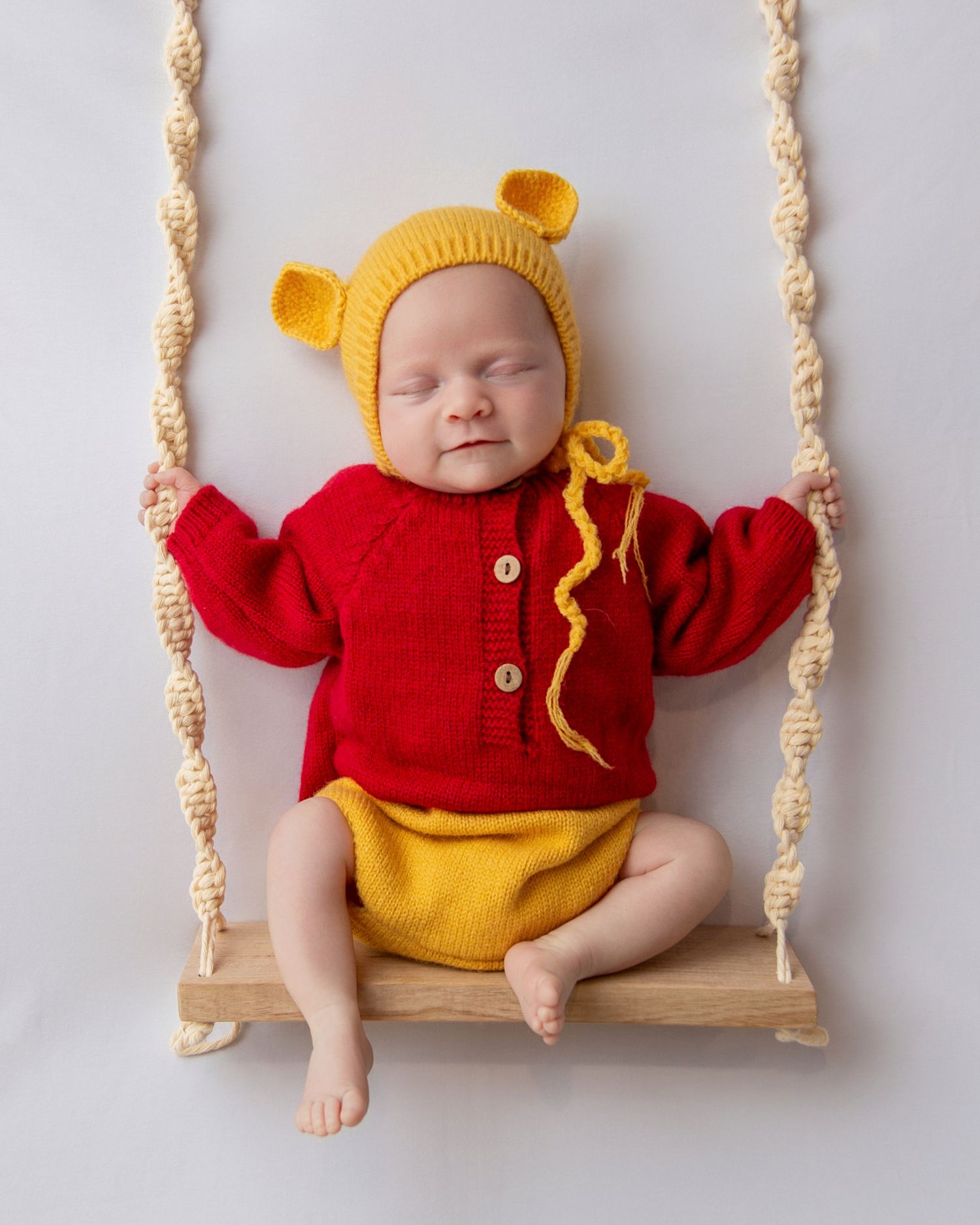 Baby wearing red sweater and yellow hat sitting on a macrame rope swing against white background.