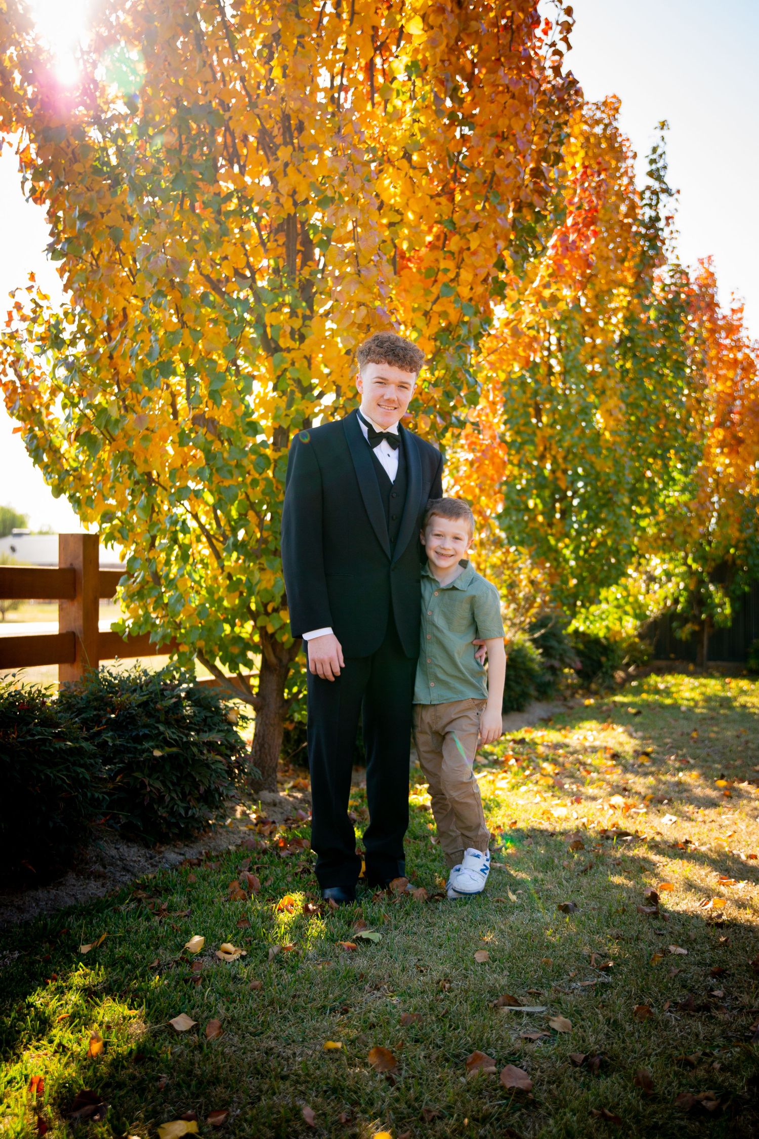 Two people stand together outdoors amidst vibrant golden autumn trees in formal attire on a sunny afternoon.