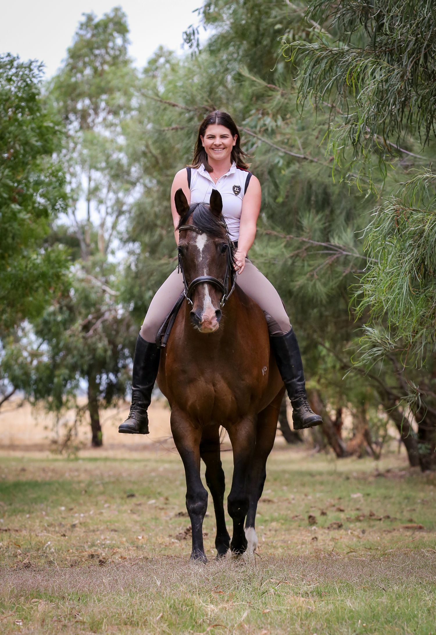 An equestrian rides a brown horse through a tree-lined path in casual attire.