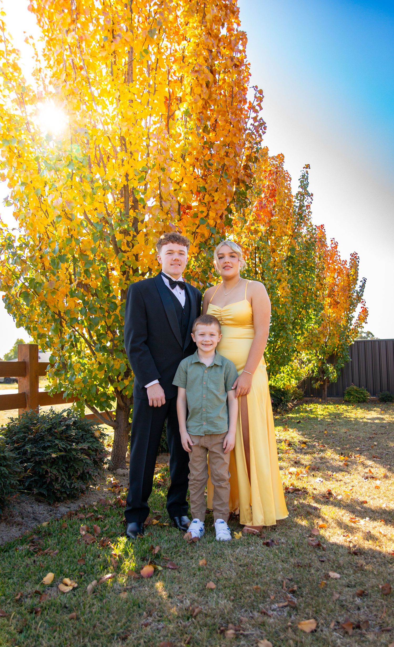 Family poses together for formal portrait in autumn with vibrant yellow trees and sunlight in background.