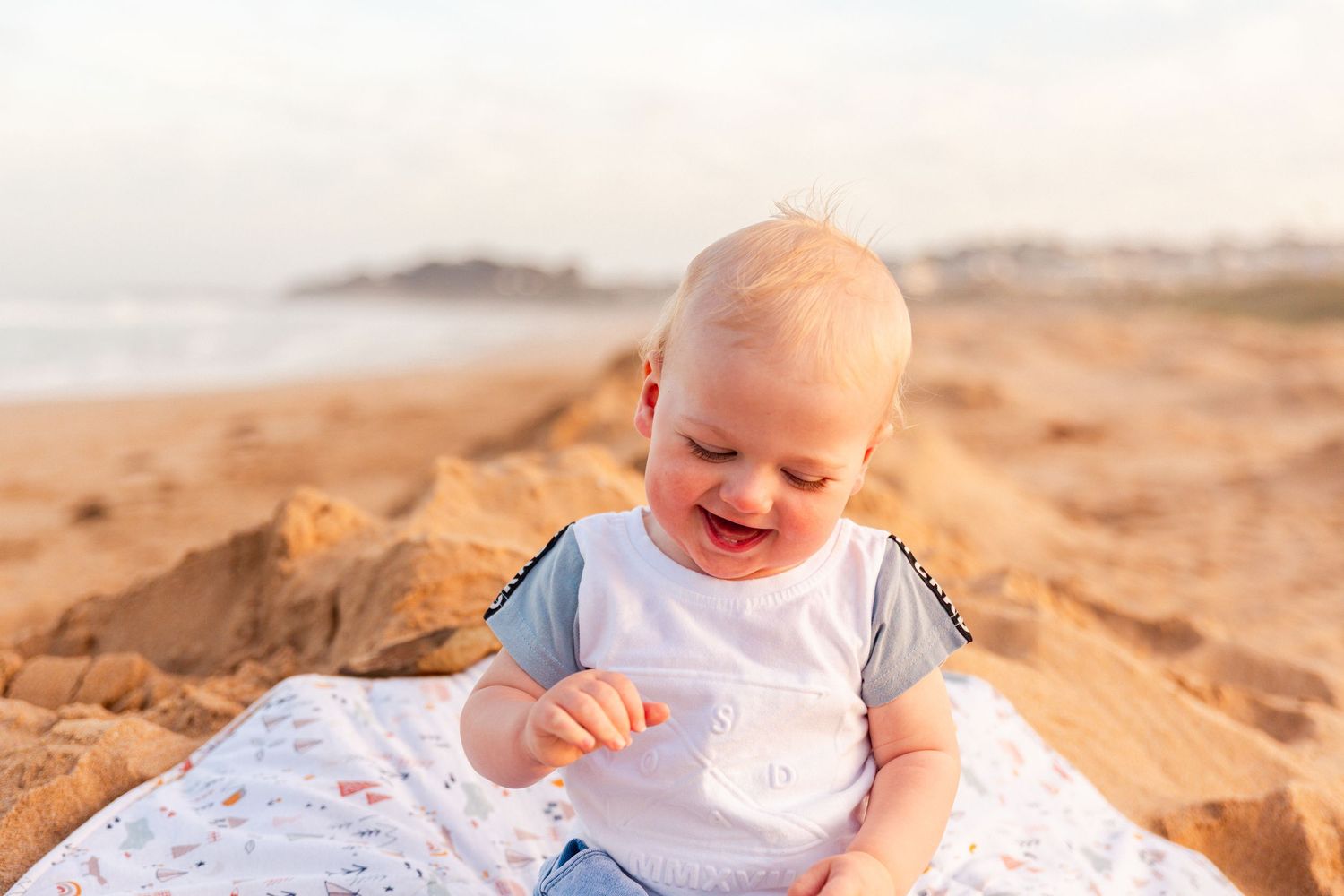 A happy baby enjoys a sunny beach day while sitting on a white blanket near coastal rocks.