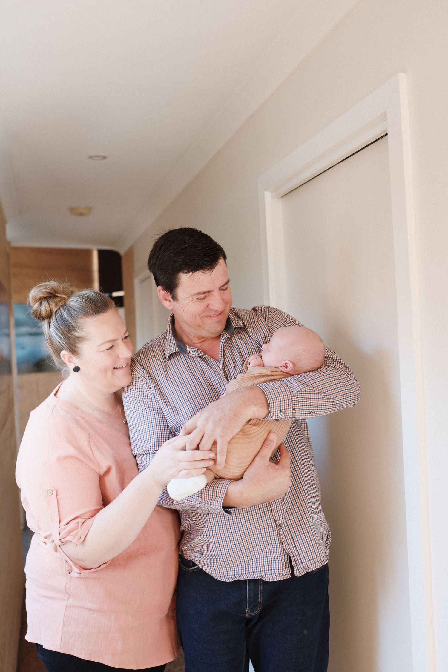 Intimate indoor family moment with parents cradling their newborn baby against a white wall.