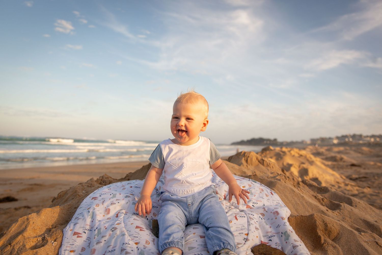 A joyful toddler sits on a blanket at the beach during golden hour with ocean waves in the background.