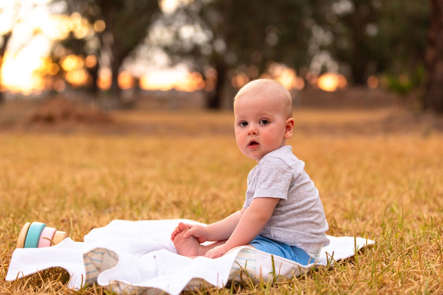 Baby in gray shirt sitting on white blanket in golden sunlit field.