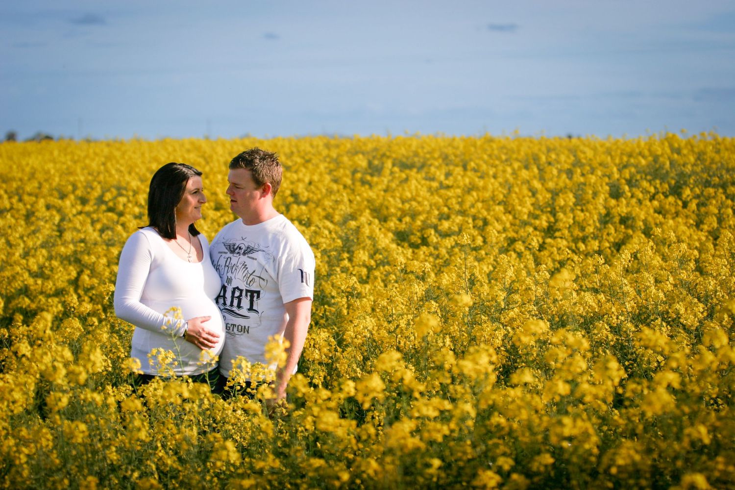 A romantic couple stands together in a vibrant yellow canola flower field against a blue sky.