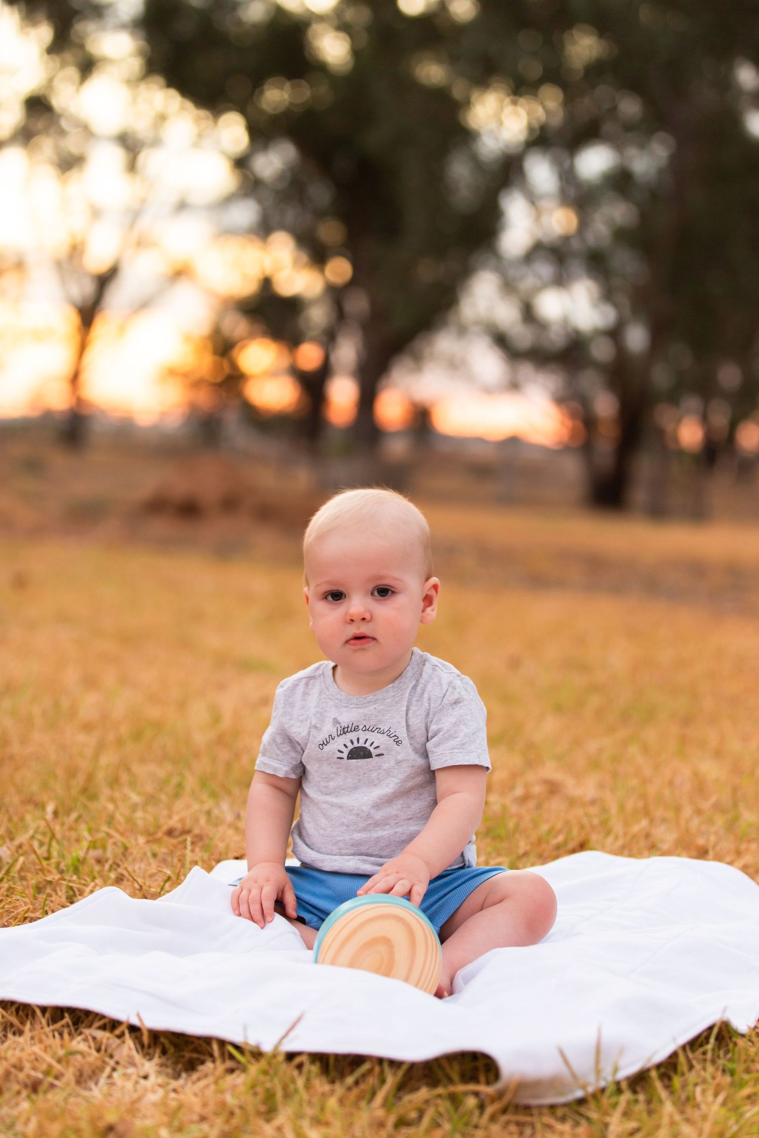 Baby in gray t-shirt sitting on white blanket in grassy field at sunset.