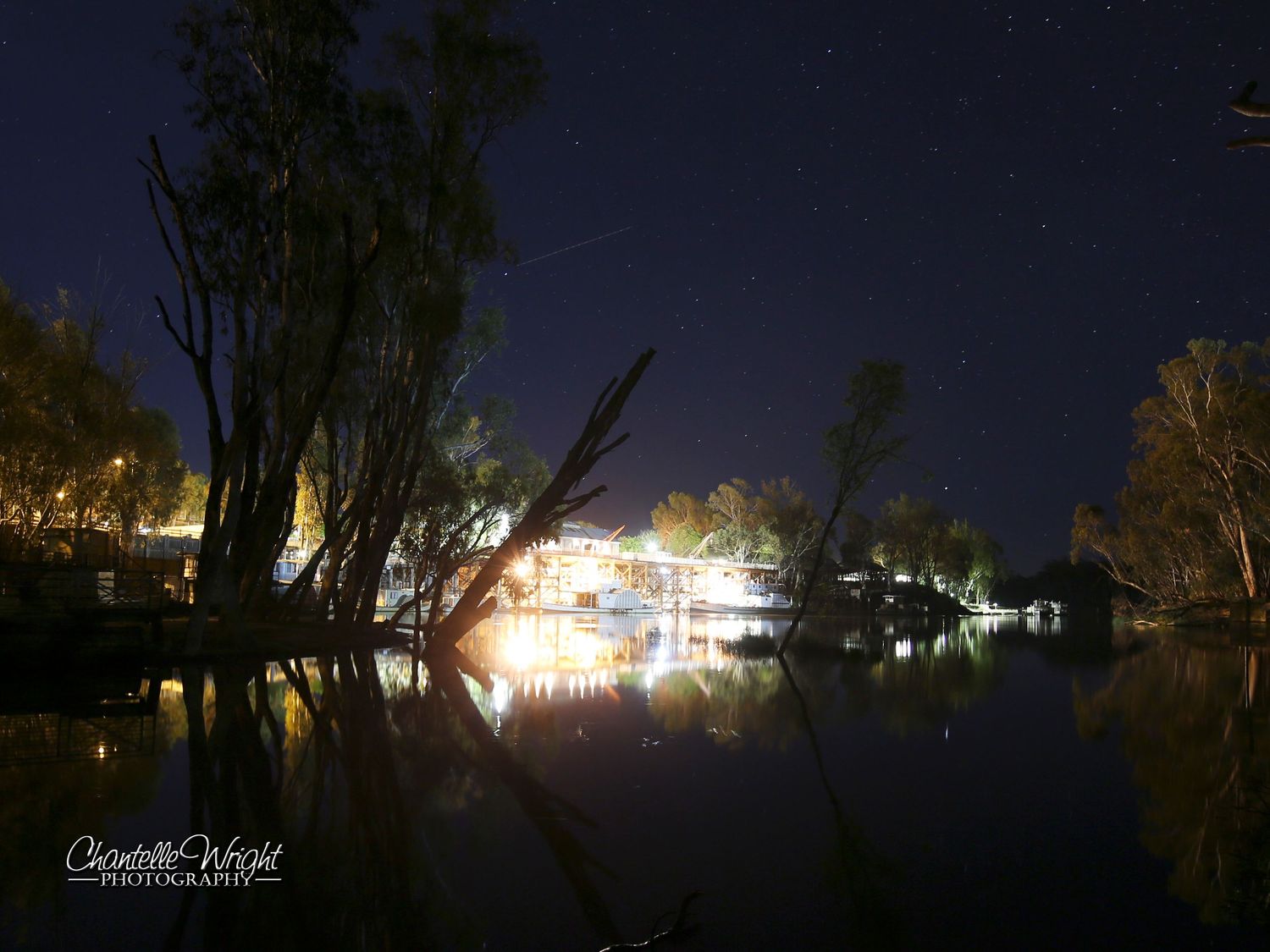 Nighttime reflection of lights on water with silhouetted trees in foreground.