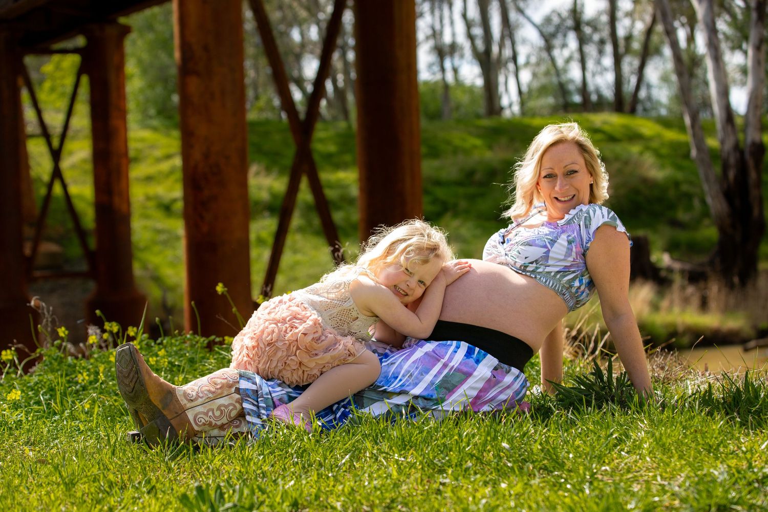 Mother and child relax together on grass near rustic structure.