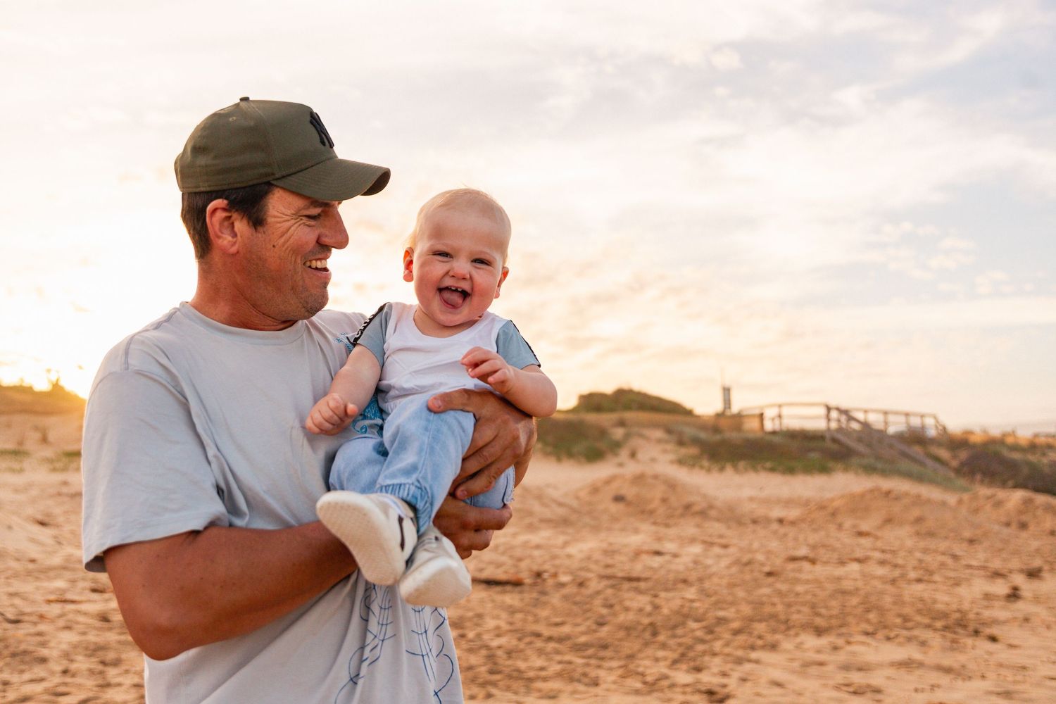 Parent and baby share laughter together in an outdoor sandy setting at sunset.