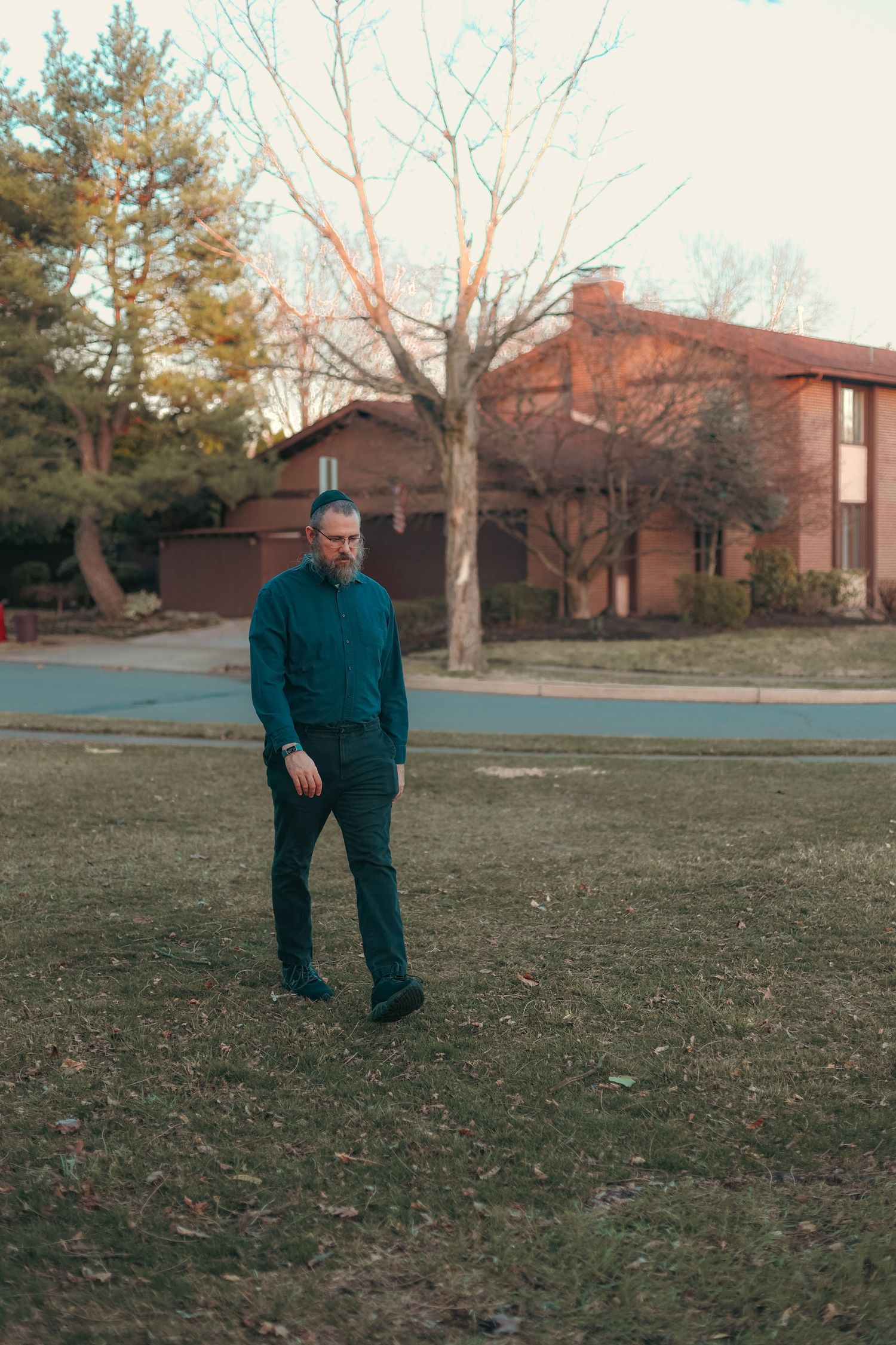 Man with beard in teal shirt walking on green lawn near brick building at sunset.