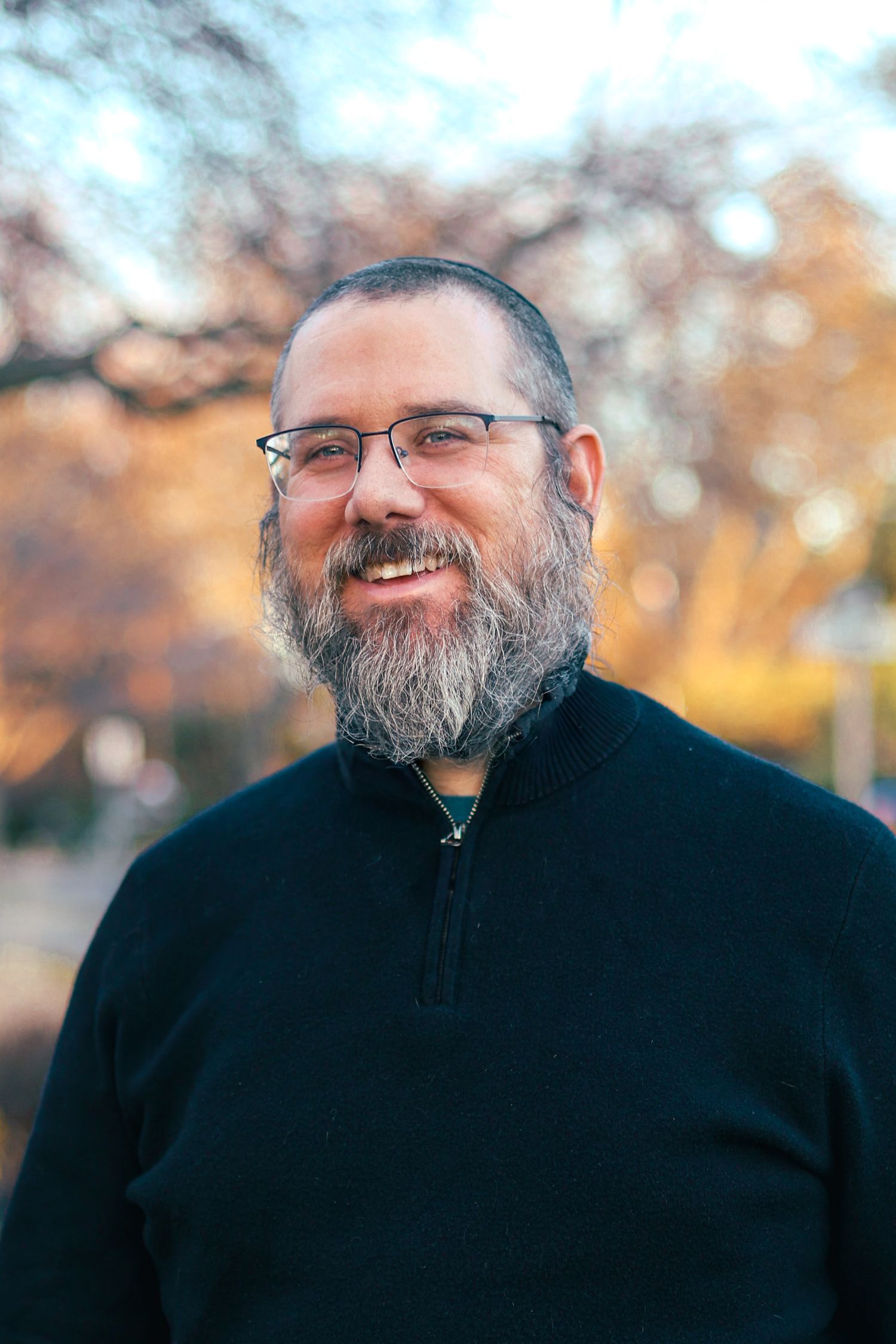 Smiling bearded man with glasses wearing a black zip-up sweater outdoors in autumn.