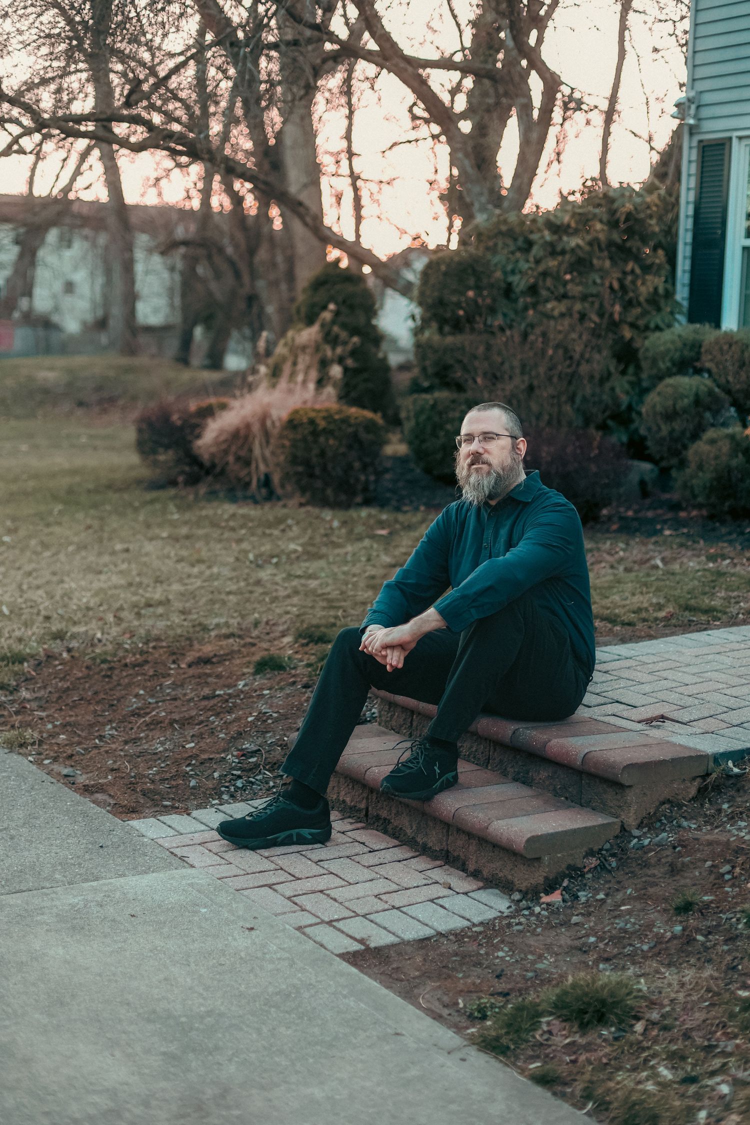 Bearded man wearing glasses sits on brick steps outside a suburban home at dusk.