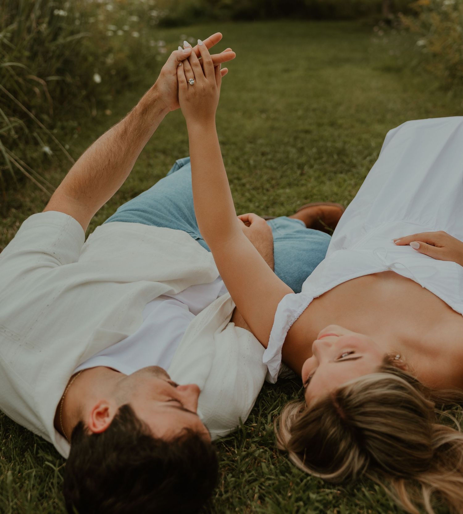 Couple lying on green grass holding hands, wearing white and blue outfits, in a romantic outdoor engagement photo.