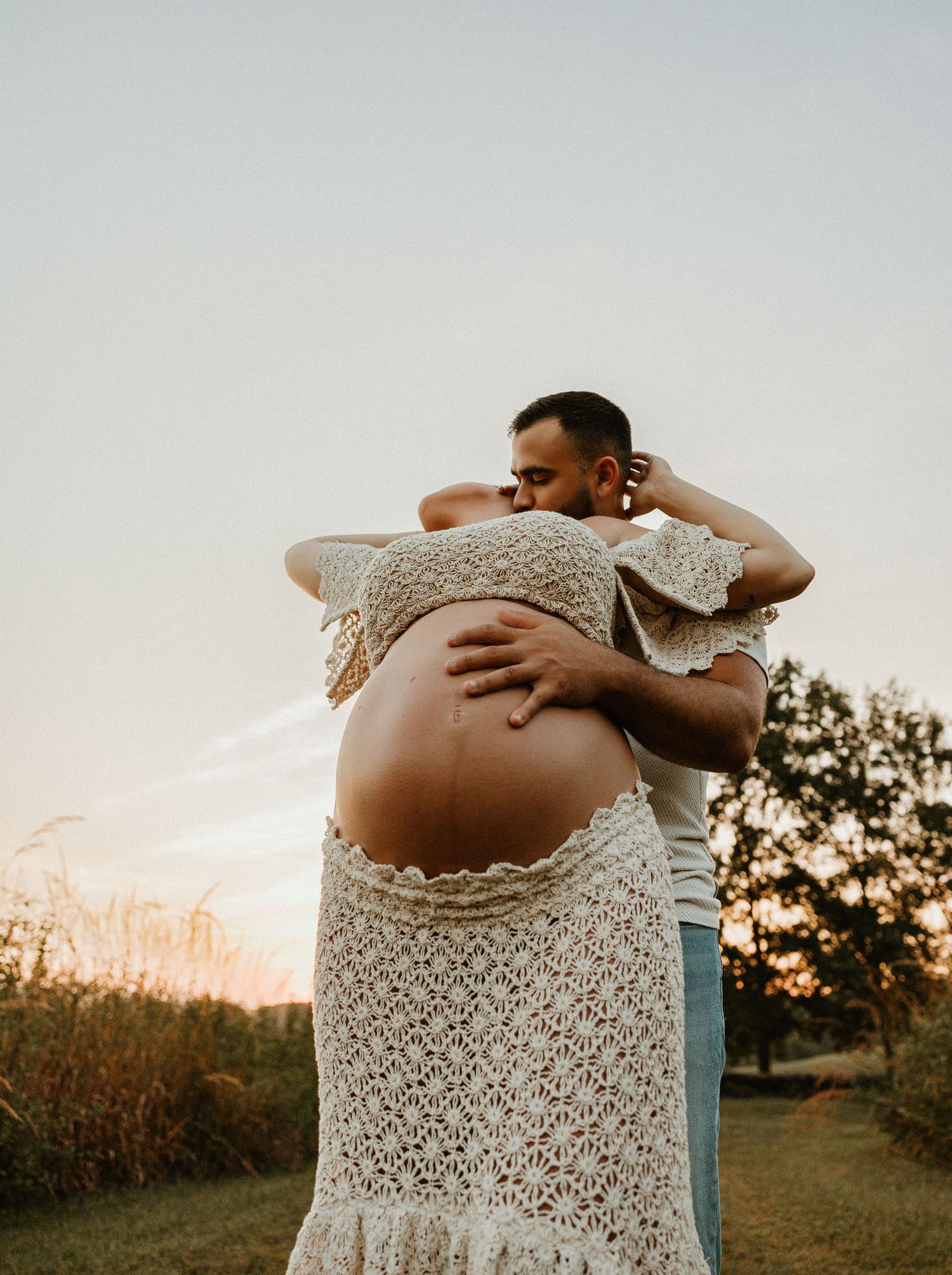 A tender maternity photoshoot moment captured at sunset in a field with a white lace dress.