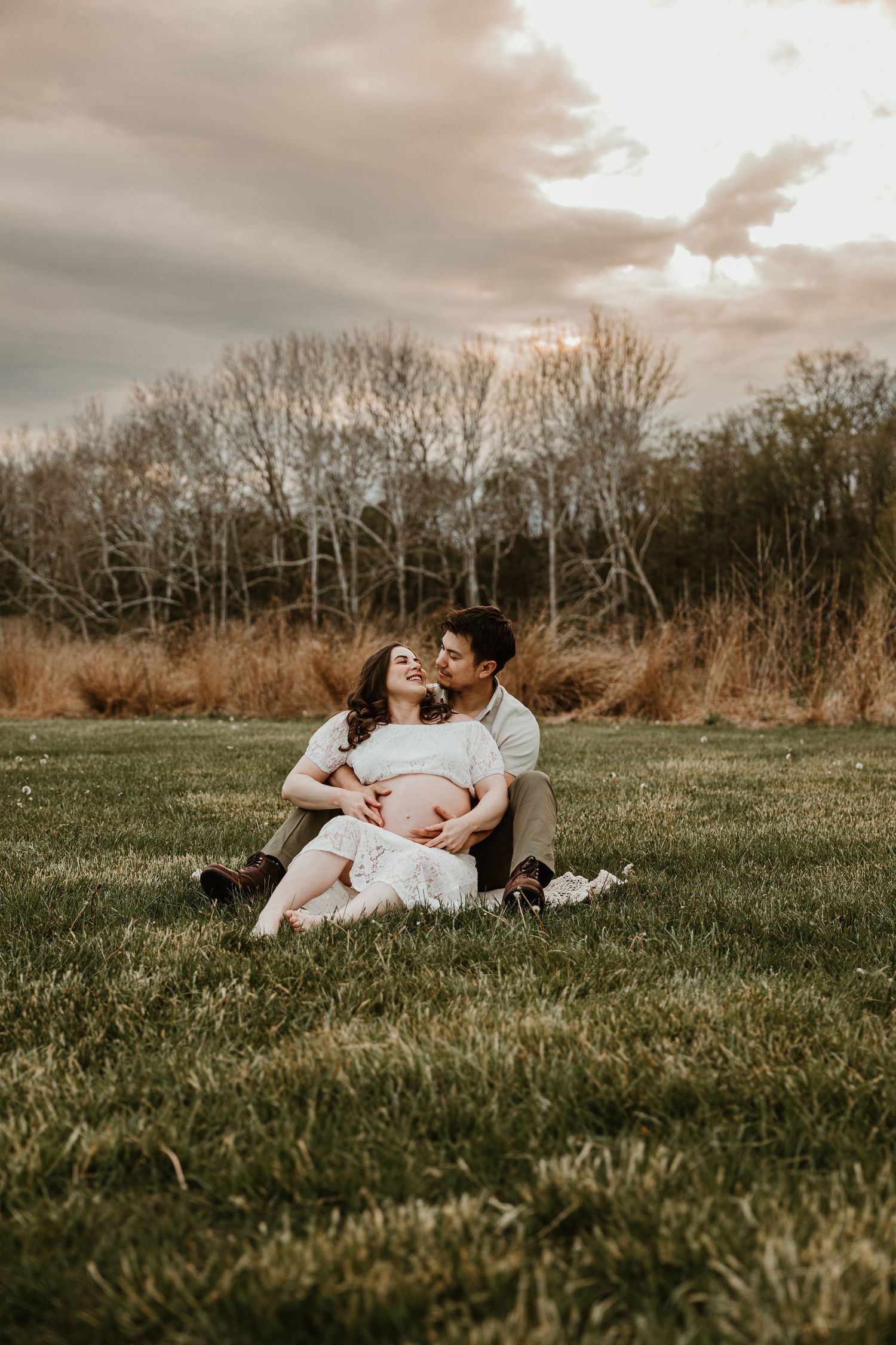 Couple sitting on grass at sunset, man embracing pregnant woman from behind in outdoor maternity photo.