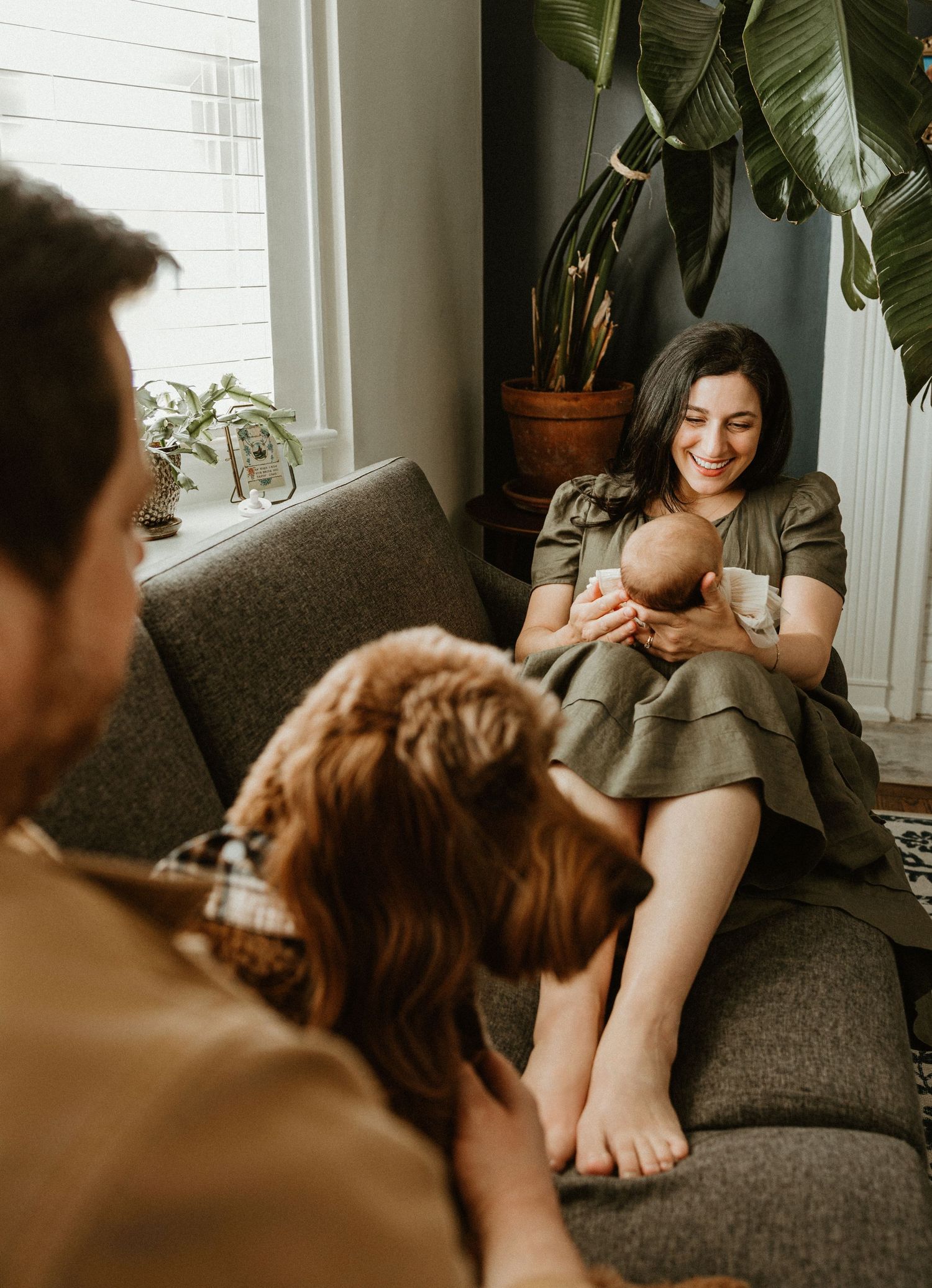 A smiling mother holds her newborn baby on a gray sofa while a man and child look on in a cozy living room.